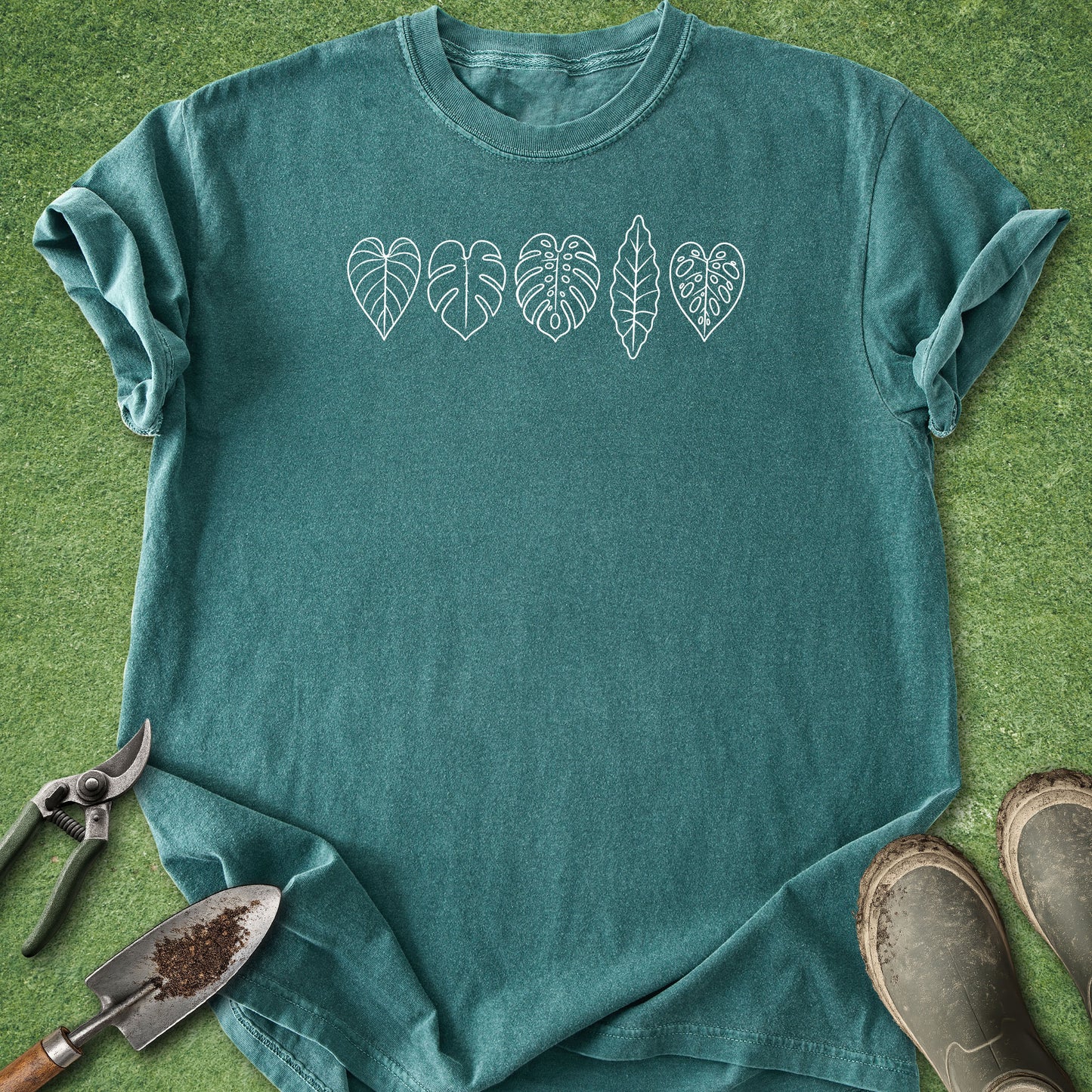 A teal short-sleeve t-shirt with a white line-art graphic of five different leaves across the chest, styled flat on green grass with gardening tools and boots.