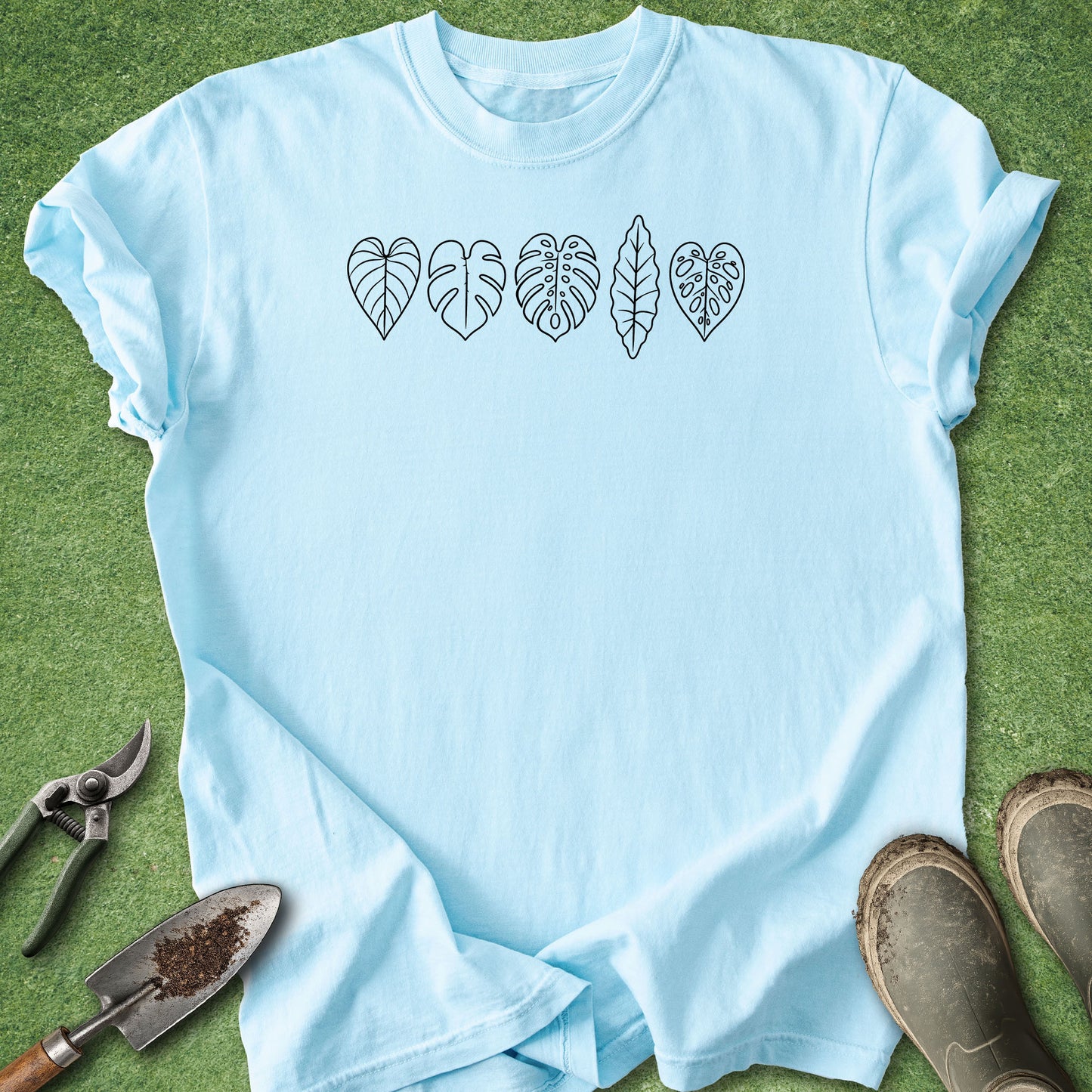 A light blue short-sleeve t-shirt laid flat on green grass, featuring a black line-art graphic of five different tropical leaves across the chest. 