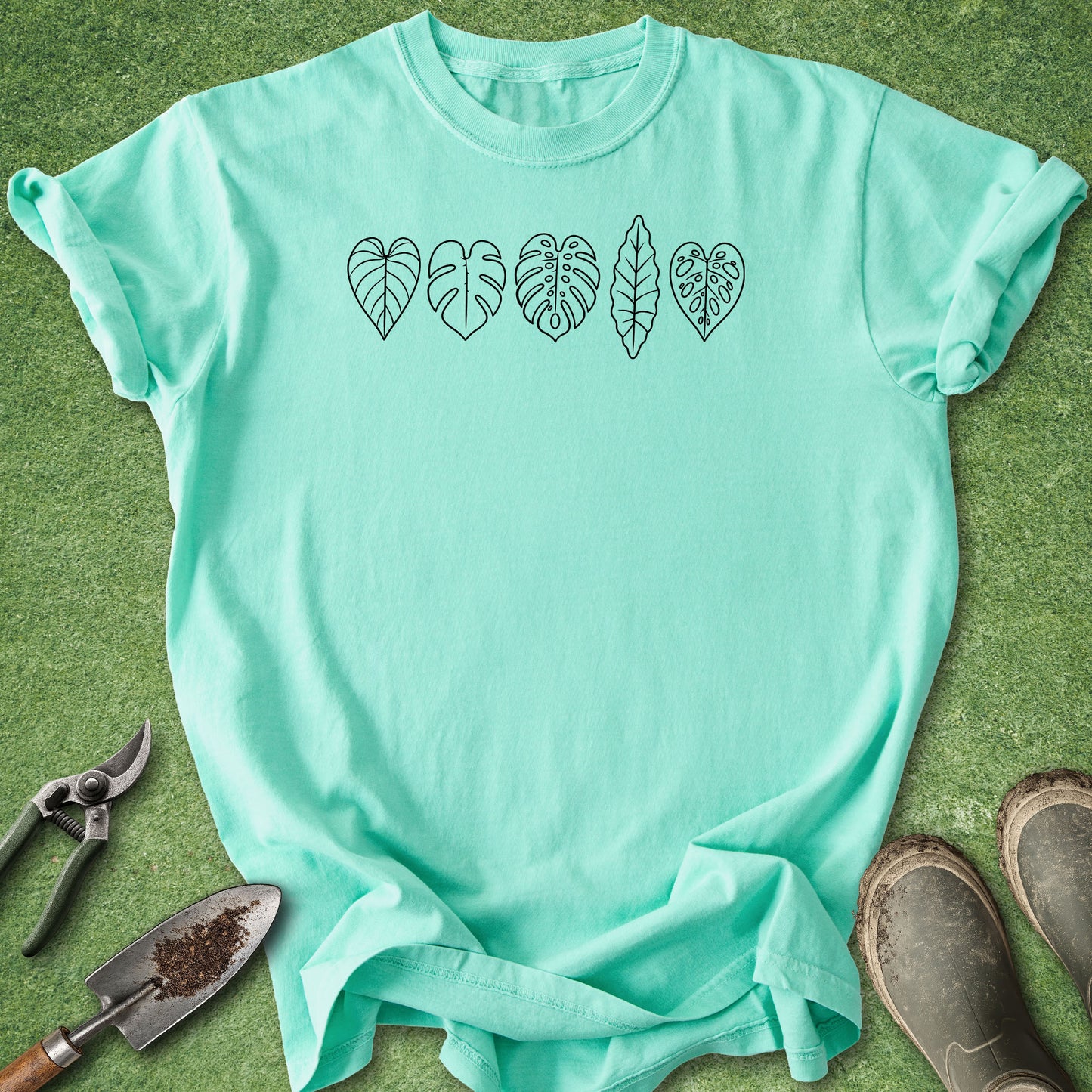 A mint green short-sleeve t-shirt with a black line art graphic of five different monstera and philodendron leaves across the chest, laid flat on green grass with gardening tools (trowel, pruning shears) and work boots nearby.