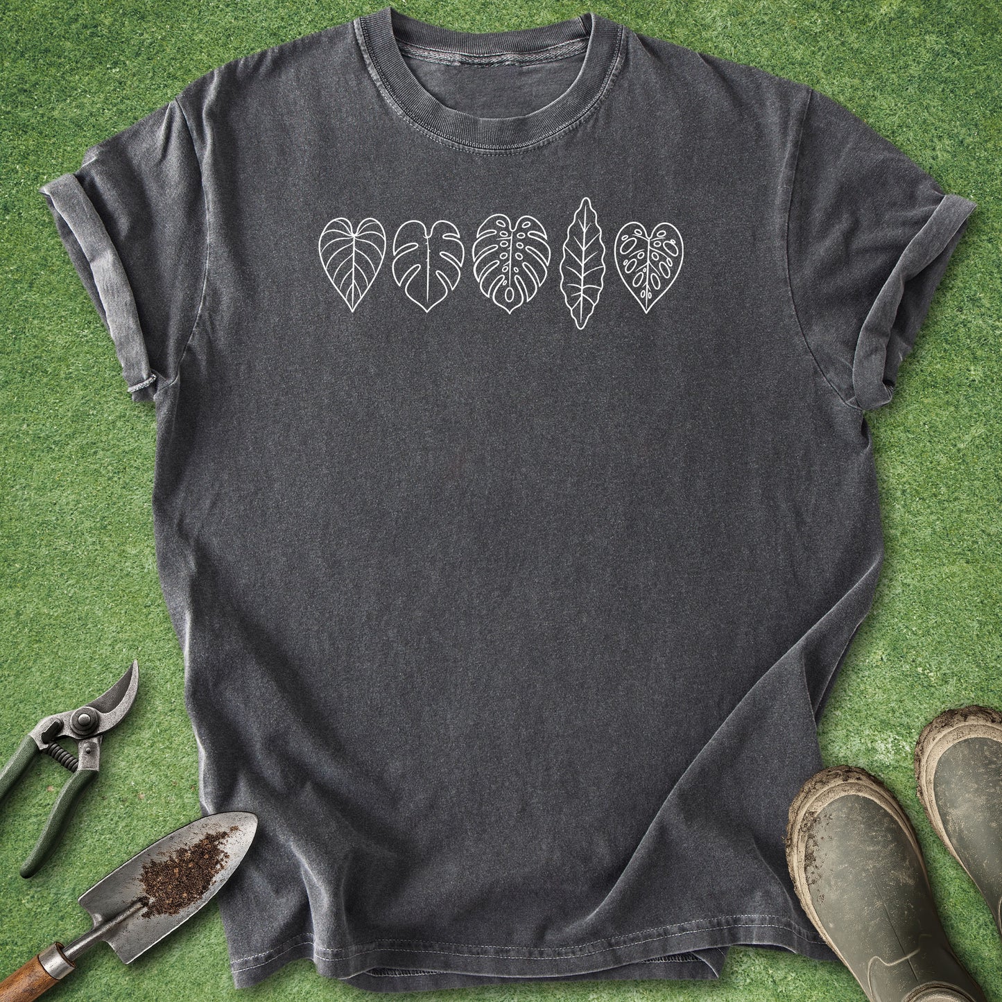 A dark charcoal gray t-shirt with five white leaf graphics across the chest, laid flat on green grass with gardening tools and boots nearby.
