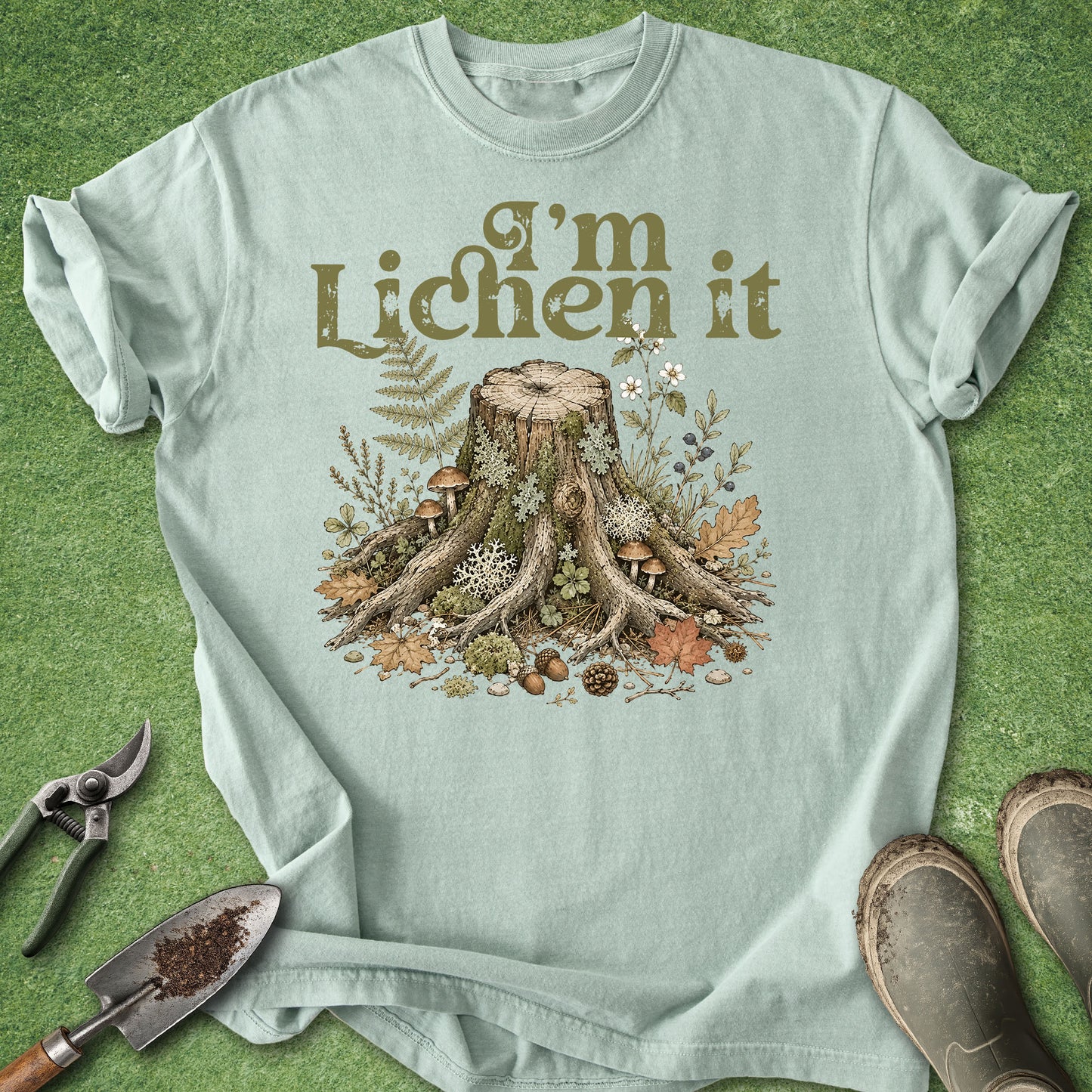 A light sage green t-shirt laid flat on a green grass-like surface, featuring a graphic of a tree stump with mushrooms and the text 'I'm Lichen it' in olive green. Gardening props including a trowel, pruning shears, and boots are visible around the shirt.