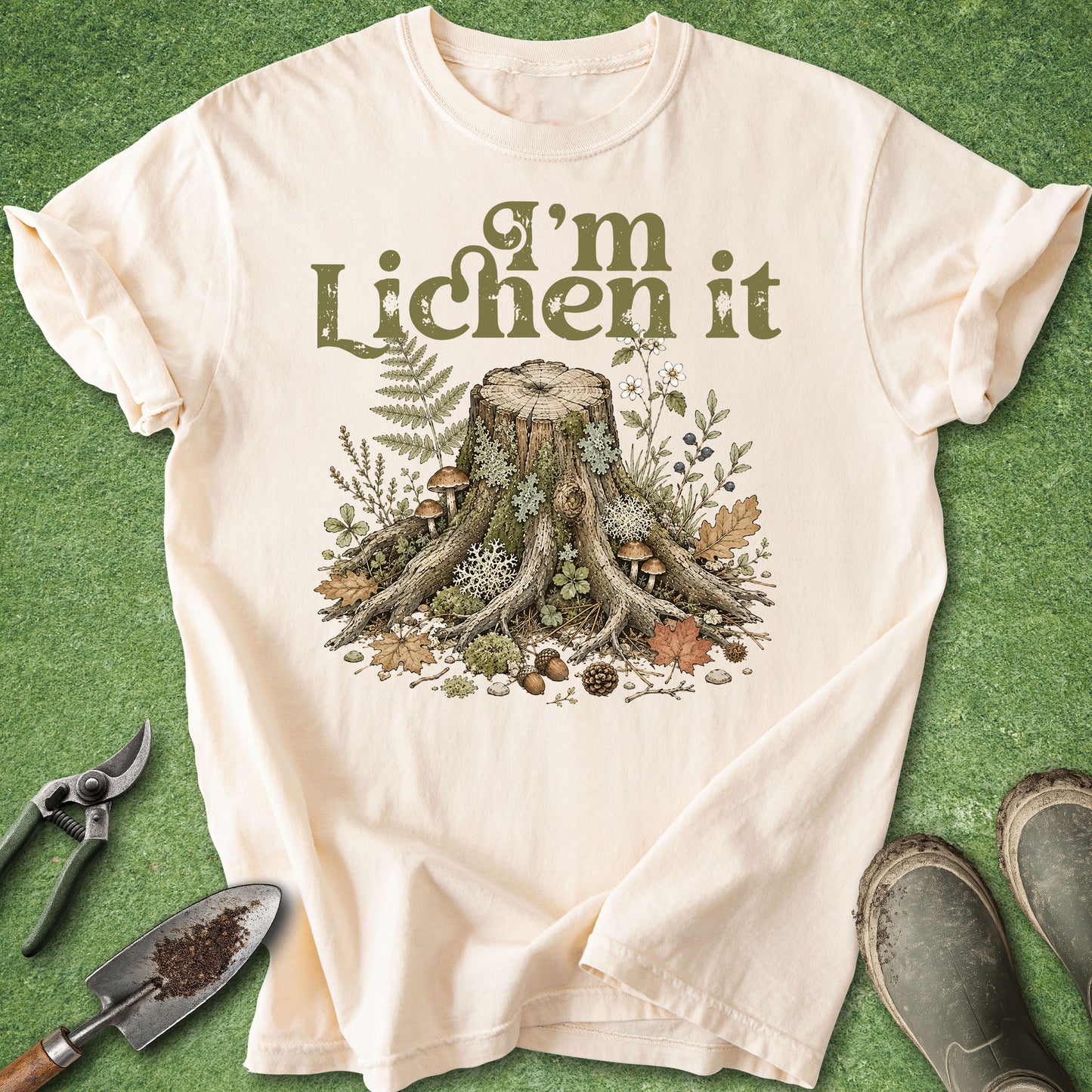 A cream-colored short-sleeve t-shirt with a graphic of a tree stump surrounded by lichen, moss, and small mushrooms, with the text 'I'm Lichen it' printed above. The shirt is laid flat on a green grass-like background with gardening props (trowel, pruning shears, boots) around it.