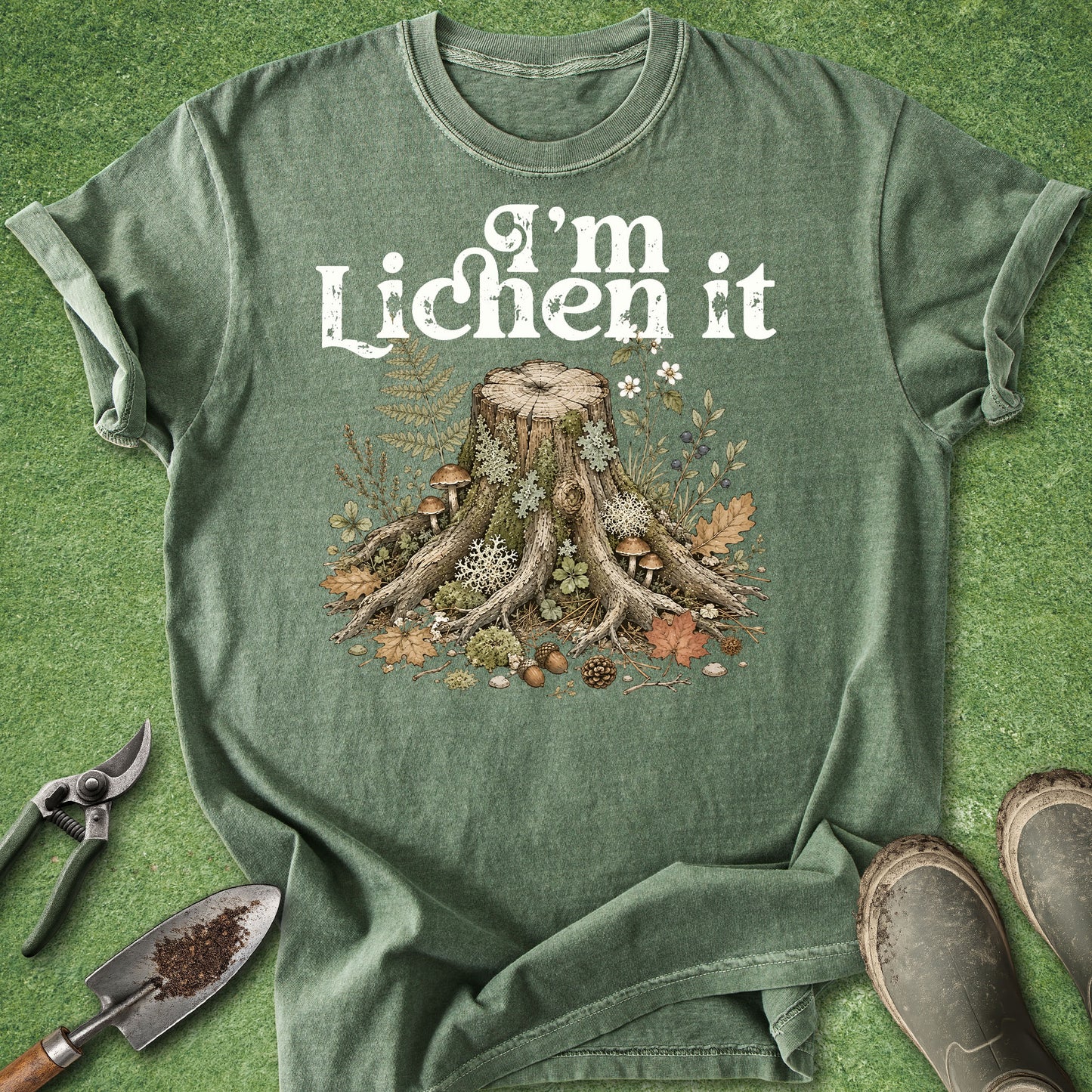 A green graphic t-shirt laid flat on a green grass-like surface, featuring a white 'I'm Lichen it' text above a detailed forest tree stump illustration with roots, mushrooms, leaves, and flowers. Gardening props including pruning shears, a trowel, and boots are placed around the shirt.