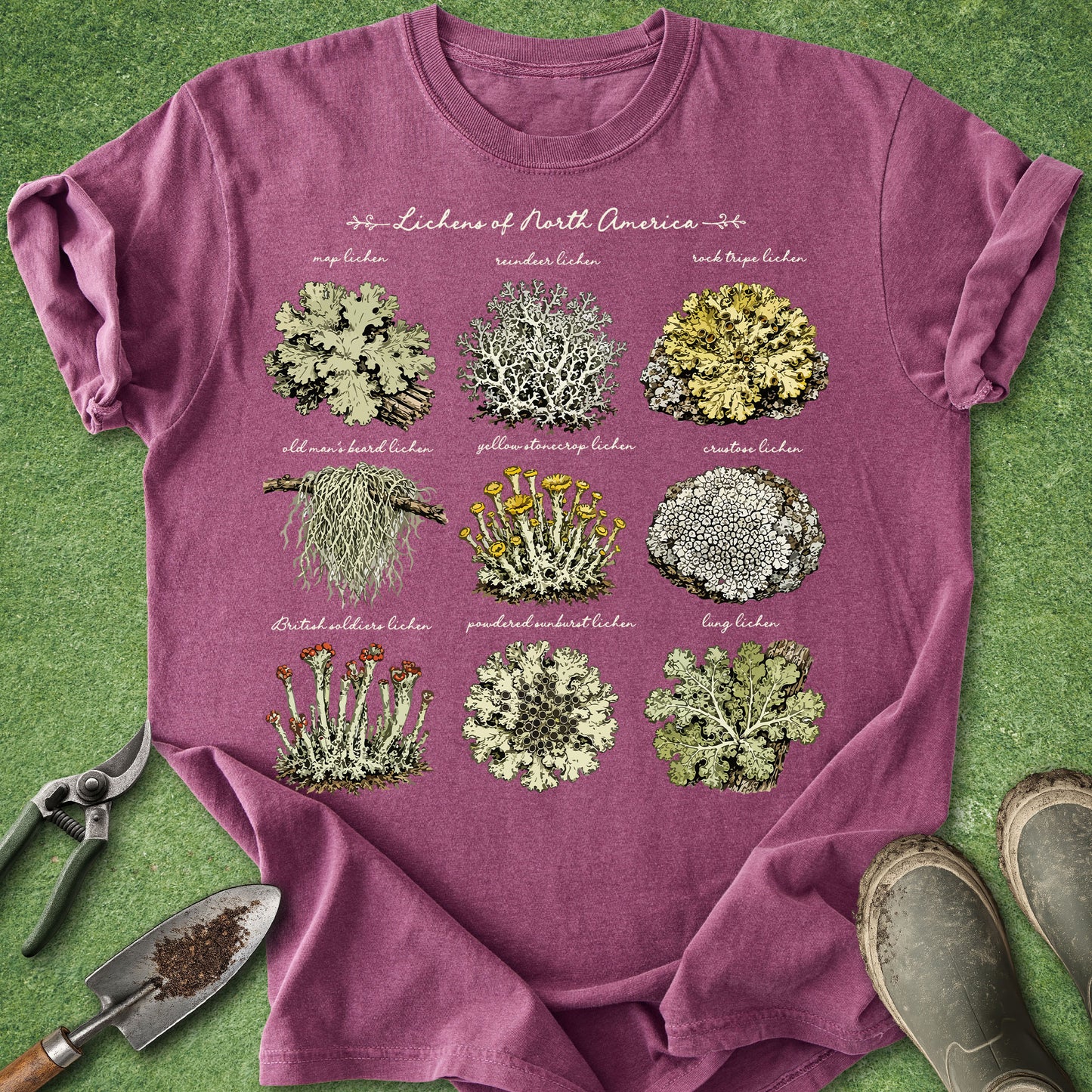 A heathered mauve-purple short-sleeve t-shirt laid flat on a green grass-like surface, featuring a large botanical graphic titled 'Lichens of North America' with nine illustrated lichen samples and small labels. Gardening props including pruning shears, a trowel, and boots are visible around the shirt.