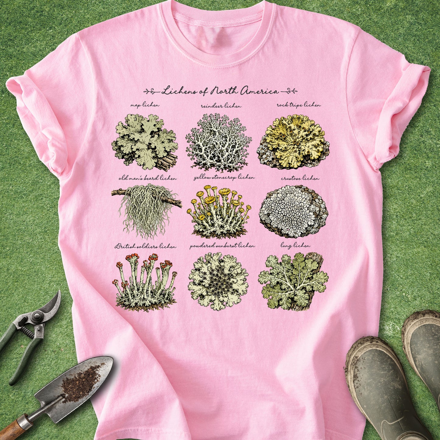 A pink short-sleeve graphic T-shirt laid flat on green grass, featuring a botanical print titled 'Lichen of North America' with nine illustrated lichen samples and small labels. Gardening props including pruning shears, a trowel, and boots are placed around the shirt.