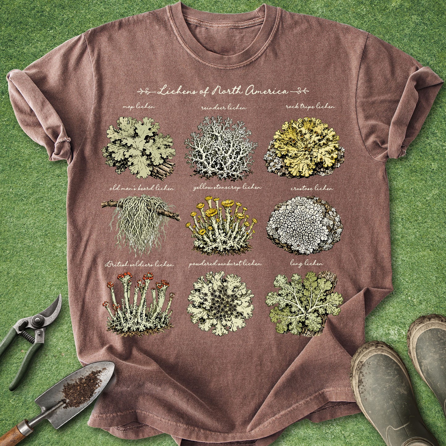 A brown short-sleeve graphic T-shirt laid flat on green grass-like background, featuring a botanical print of North American lichens with labeled illustrations. Gardening props including pruning shears, a small trowel, and boots are placed around the shirt.