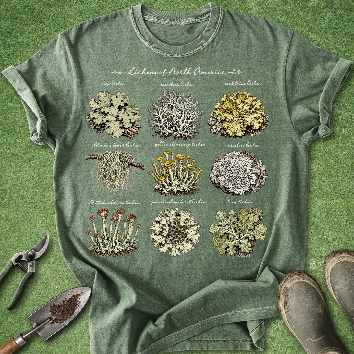 An olive green graphic t-shirt laid flat on a green grass-like surface, featuring a vintage-style print of nine different lichens labeled 'Lichen of North America.' Gardening props including pruning shears, a trowel, and boots are placed around the shirt.