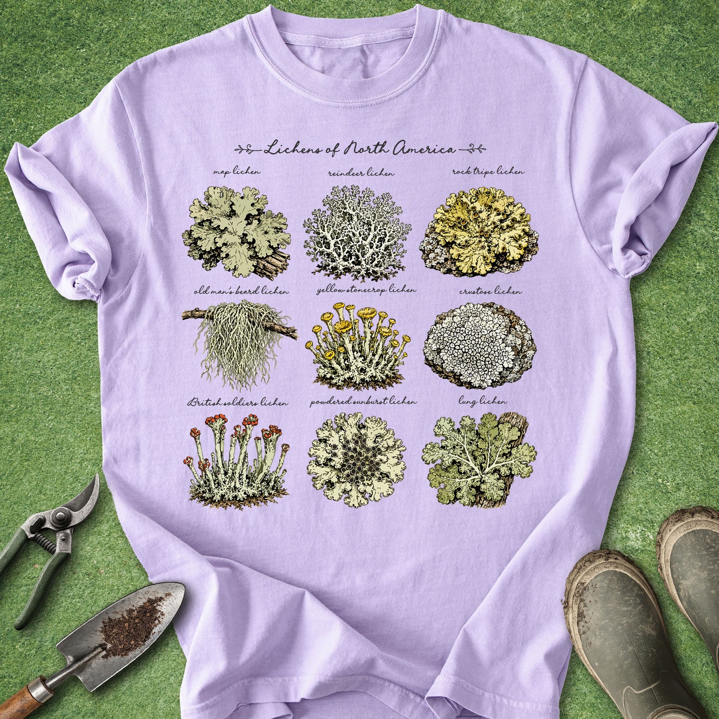 A light lavender crewneck t-shirt featuring a detailed botanical graphic of North American lichens, styled in a flat-lay on green grass with gardening tools and boots.