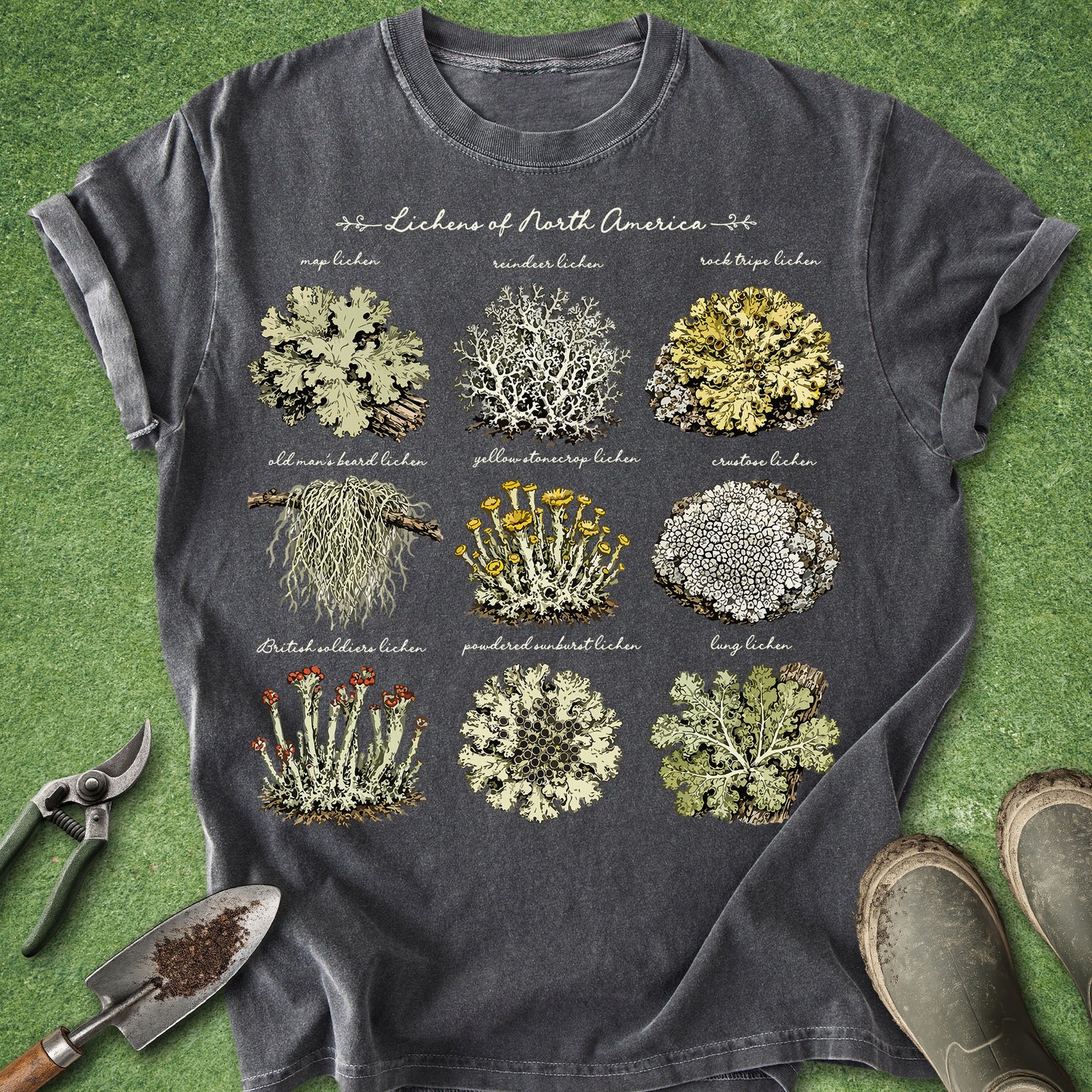 A dark gray graphic t-shirt laid flat on green grass, featuring a detailed botanical print of 'Lichen of North America' with nine different lichen species. Gardening props including pruning shears, a trowel, and boots are visible around the shirt.