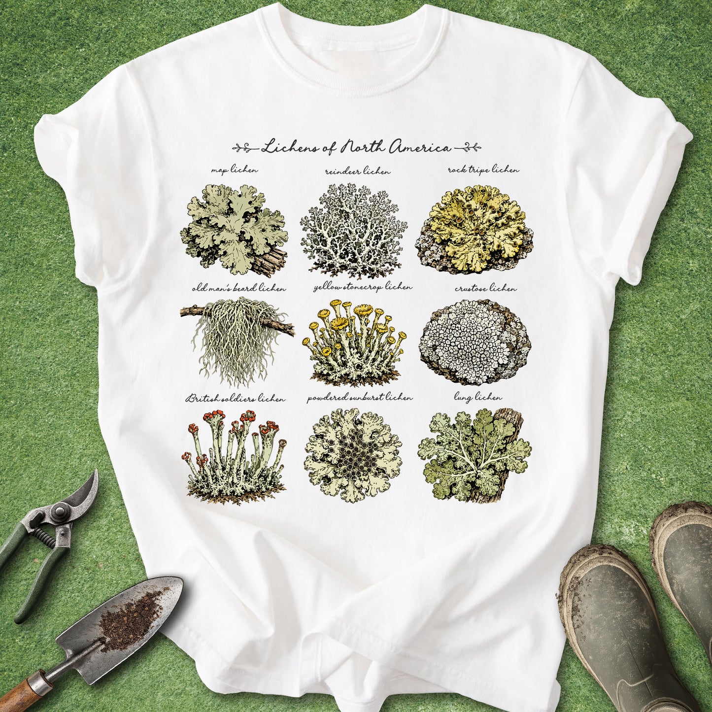 A white graphic T-shirt laid flat on a green textured surface, featuring nine illustrated patches of North American lichens with labels. The shirt is styled with gardening tools and boots around it.