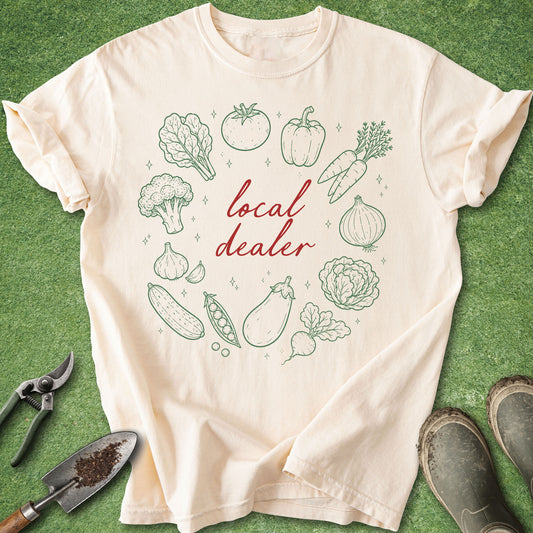 Ivory Comfort Colors t-shirt featuring the phrase "local dealer" surrounded by hand-drawn vegetables including tomatoes, peppers, carrots, onions, broccoli, garlic, cucumbers, peas, cabbage, eggplant, and leafy greens.