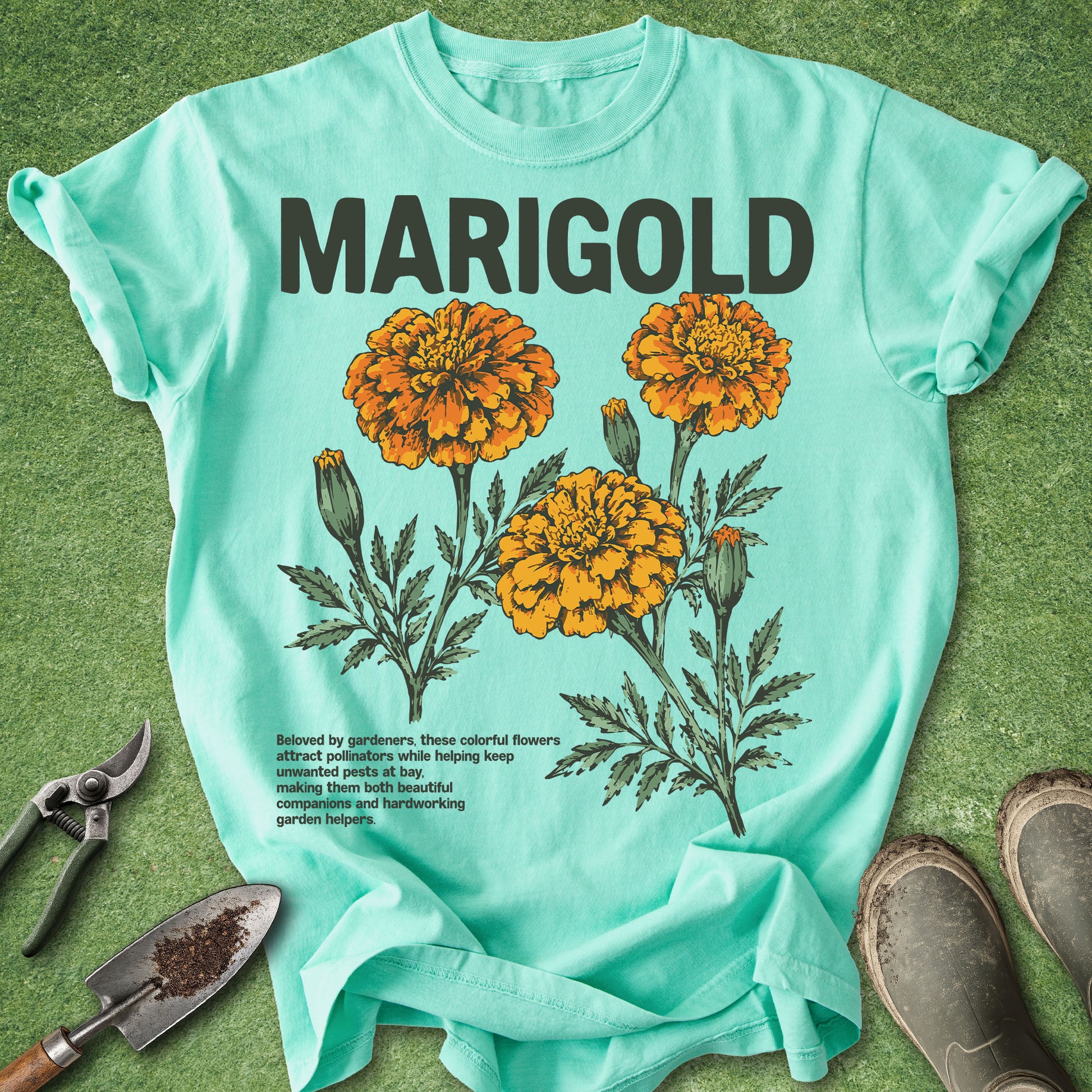 Light blue t-shirt with marigold flower design and text on a green surface with gardening tools.