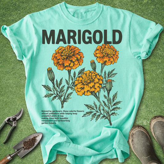Light blue t-shirt with marigold flower design and text on a green surface with gardening tools.