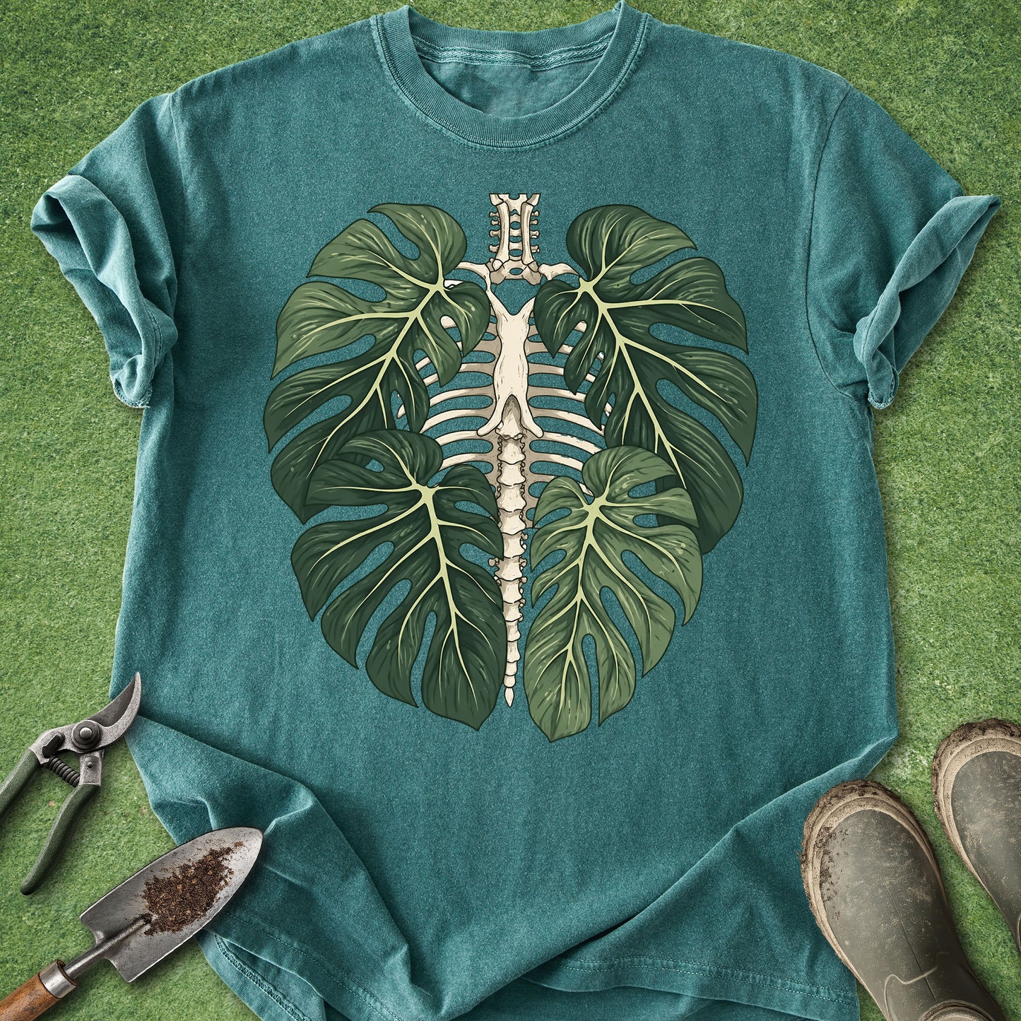 A teal short-sleeve t-shirt laid flat on green grass, featuring a large graphic of a monstera leaf pair with a white rib cage and spine running through the center. Gardening tools and boots are placed around the shirt as styling props.
