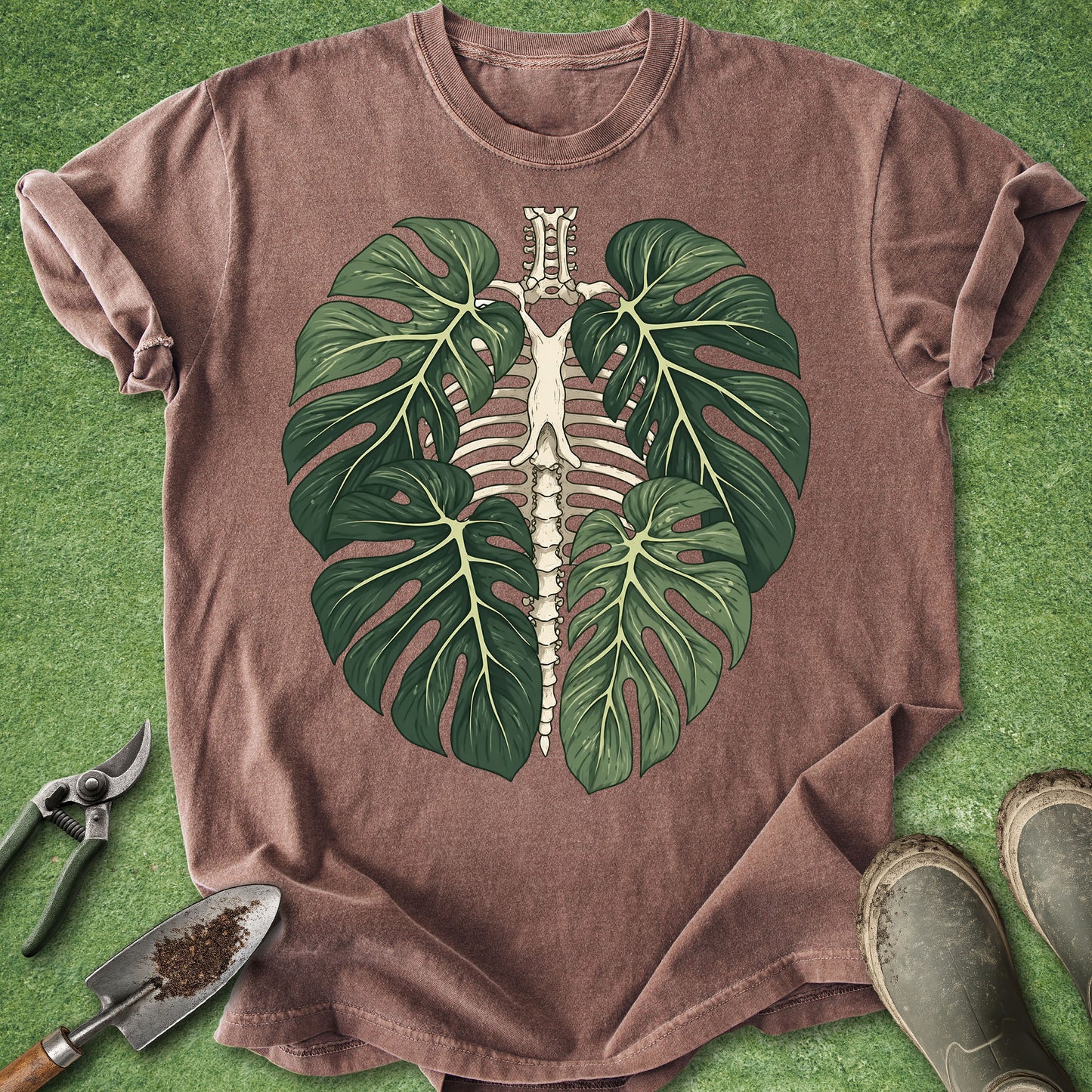 A brown short-sleeve t-shirt laid flat on green grass, featuring a graphic of a green monstera leaf skeleton. Gardening tools and boots are visible around the shirt.