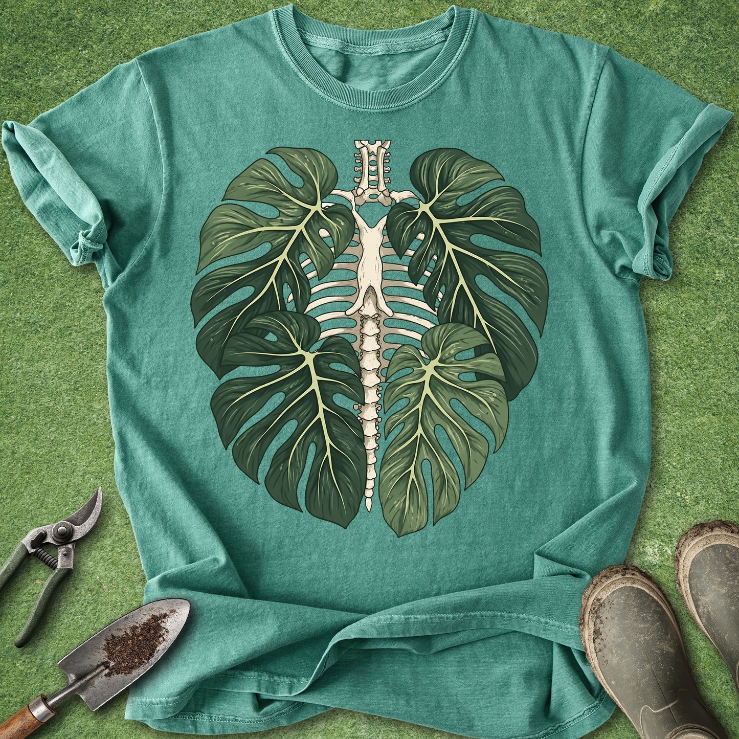 A teal short-sleeve t-shirt laid flat on a green grass-like surface, featuring a large graphic of a monstera leaf paired with a white rib cage. Gardening tools and boots are placed around the shirt as styling props.