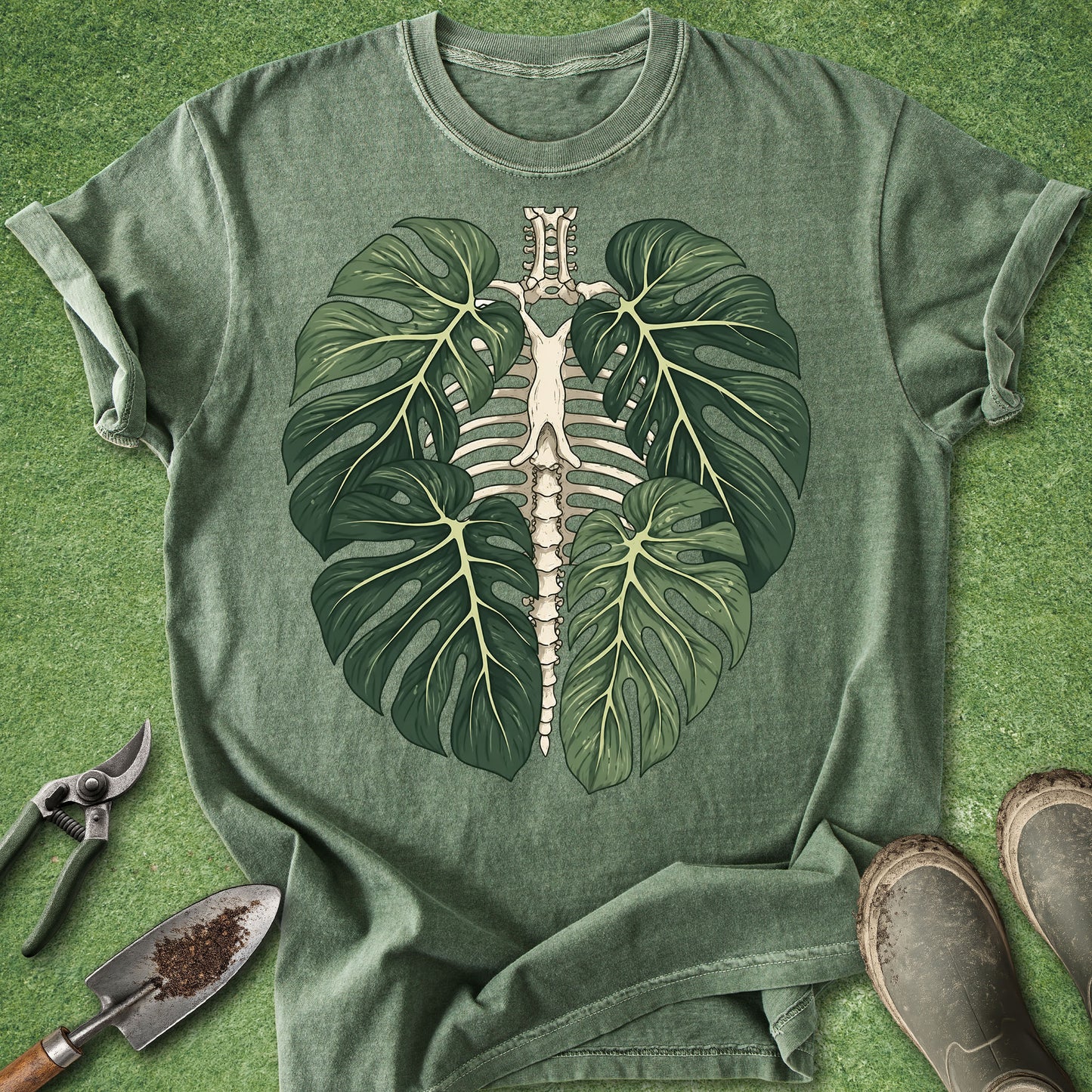 A green short-sleeve t-shirt laid flat on a green textured surface, featuring a large graphic of a human rib cage and spine framed by two green monstera leaves. Gardening tools and boots are placed around the shirt as styling props.
