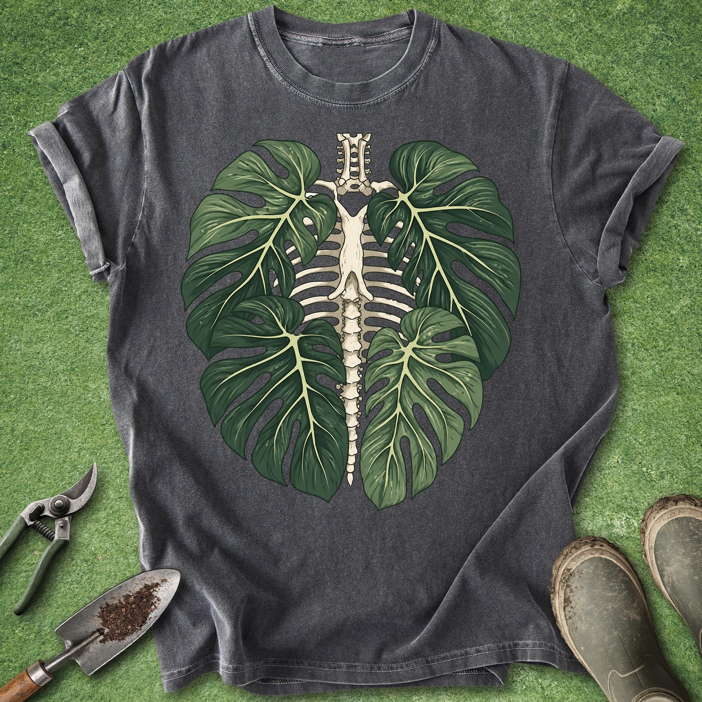 A dark charcoal gray t-shirt laid flat on green grass, featuring a graphic of a green monstera leaf skeleton. Gardening props including pruning shears, a trowel with soil, and boots are arranged around the shirt.