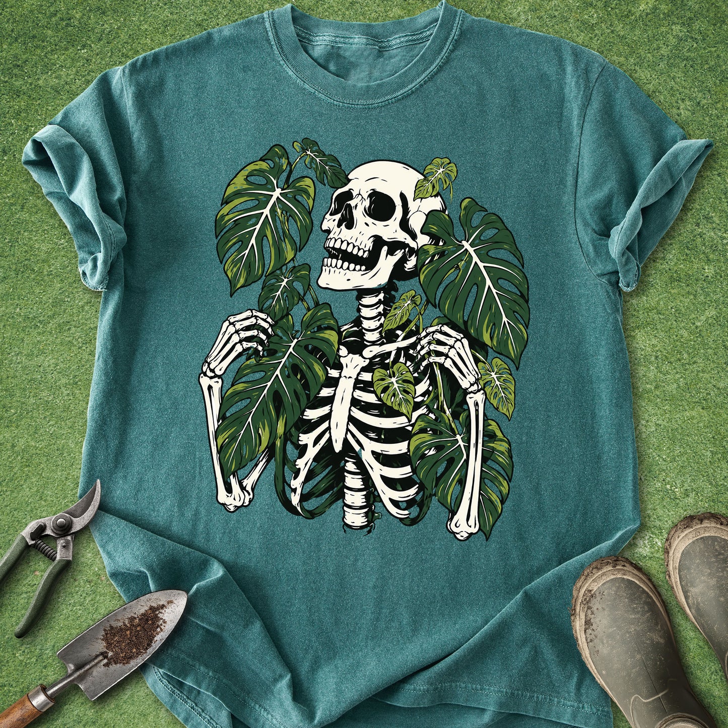 A teal short-sleeve t-shirt laid flat on green grass, featuring a graphic of a white skeleton surrounded by large green monstera leaves. Gardening tools and boots are placed around the shirt as styling props.