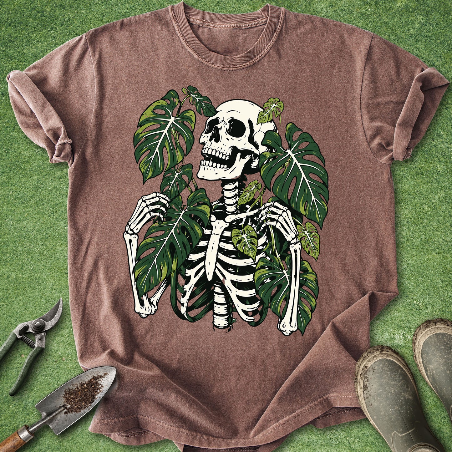 A brown short-sleeve t-shirt laid flat on a green grass-like surface, featuring a large graphic of a white skeleton surrounded by green tropical leaves. Gardening tools and boots are partially visible around the shirt as styling props.