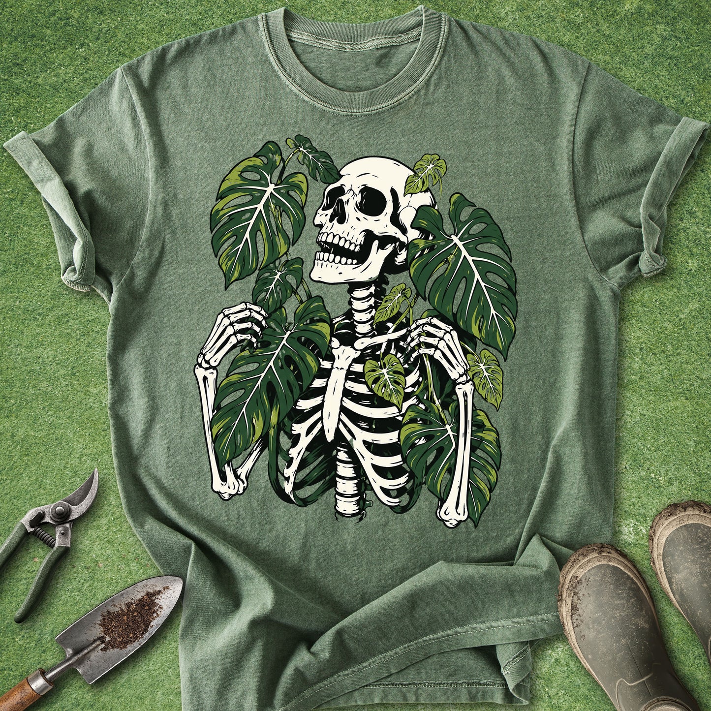 An olive green graphic t-shirt featuring a skeleton surrounded by green monstera leaves, laid flat on a green grass-like surface with gardening tools and boots nearby.