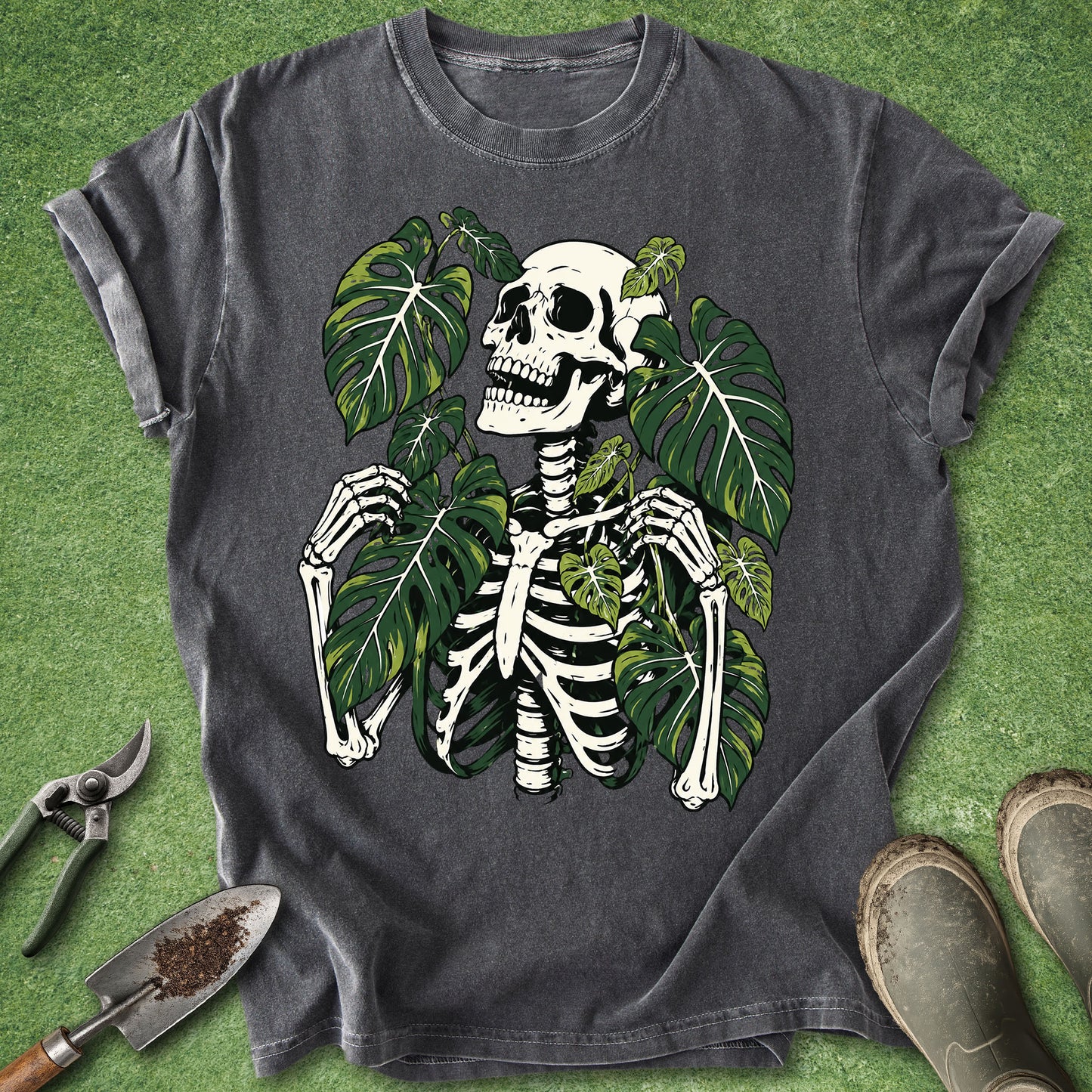 A dark gray graphic t-shirt featuring a white skeleton and green monstera leaves, laid flat on a green grass-like surface with gardening tools and boots nearby.
