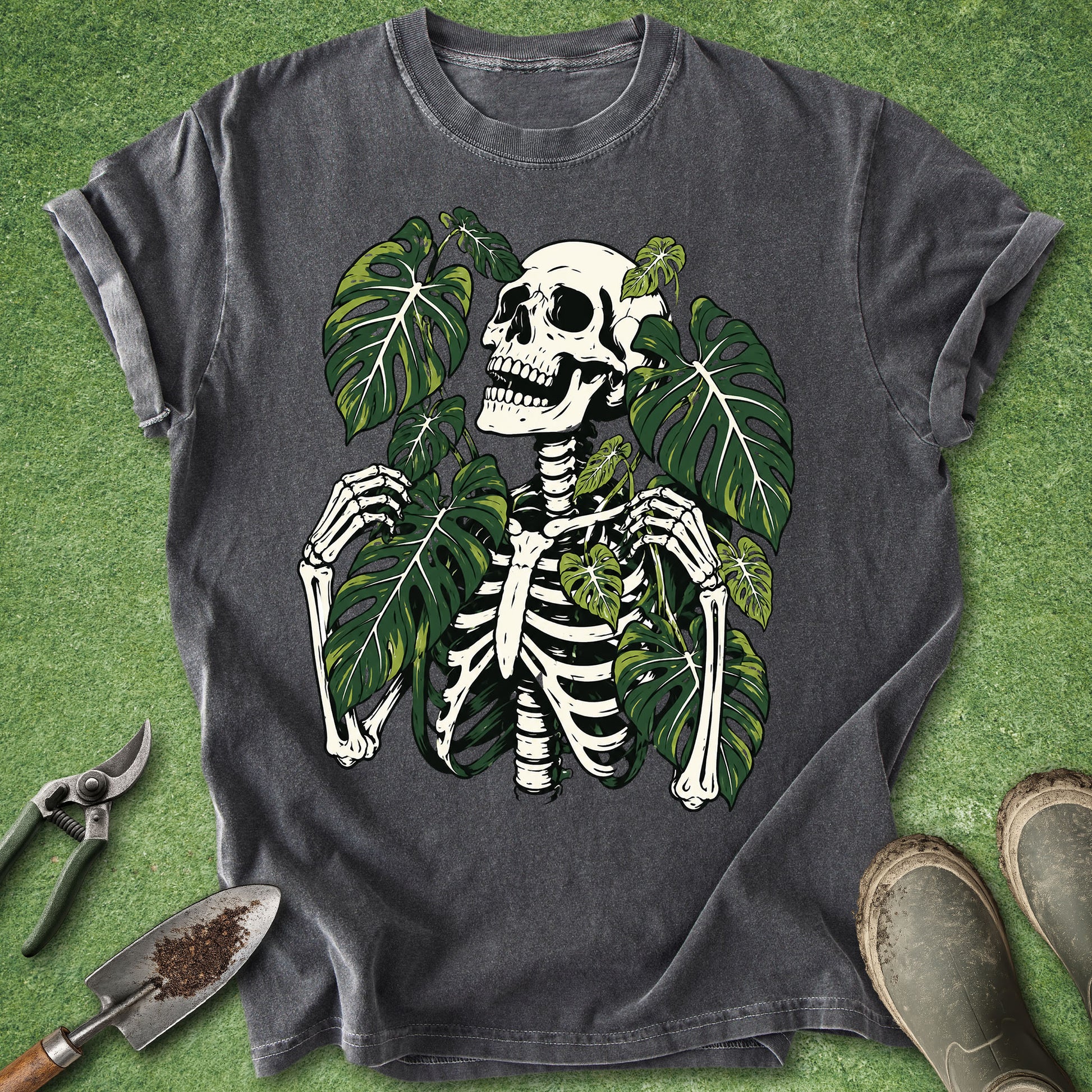 A dark gray graphic t-shirt featuring a white skeleton and green monstera leaves, laid flat on a green grass-like surface with gardening tools and boots nearby.