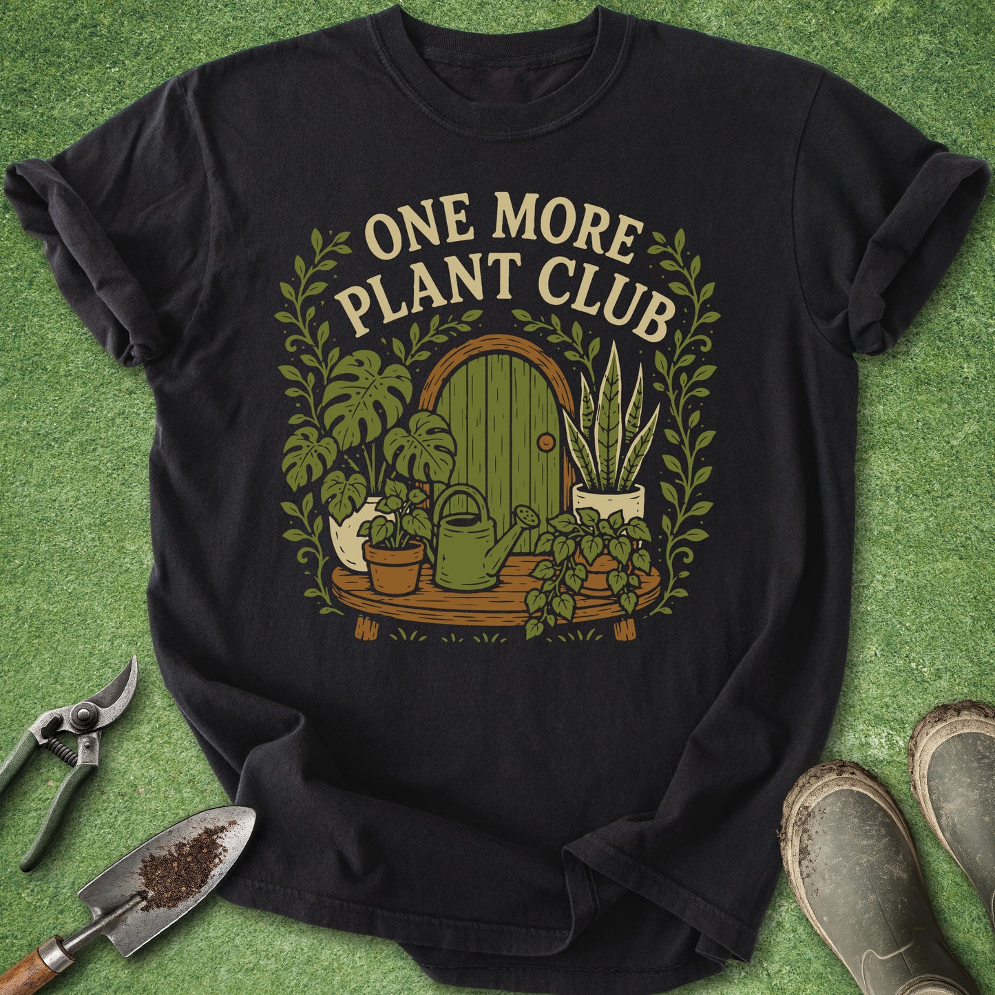 A black short-sleeve graphic T-shirt laid flat on a green grass-like surface, surrounded by gardening props including pruning shears, a trowel, and boots. The shirt features a large front print with the words “ONE MORE PLANT CLUB” above an illustrated green door, plants, pots, and vines.