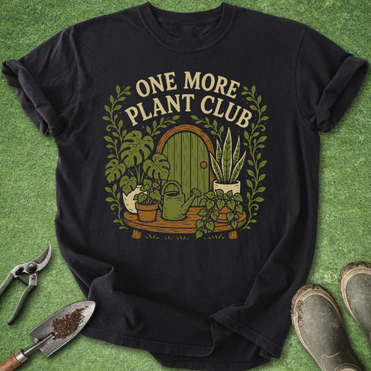A black short-sleeve graphic T-shirt laid flat on a green grass-like surface, surrounded by gardening props including pruning shears, a trowel, and boots. The shirt features a large front print with the words “ONE MORE PLANT CLUB” above an illustrated green door, plants, pots, and vines.