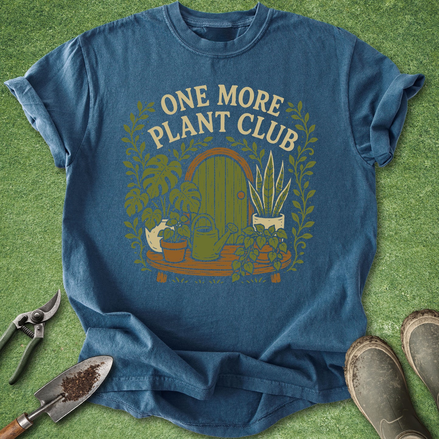 A blue graphic t-shirt laid flat on a green grass-like surface, surrounded by gardening props including pruning shears, a trowel, and boots. The shirt features a retro-style illustration of a garden door, potted plants, and the text 'ONE MORE PLANT CLUB' in cream lettering.