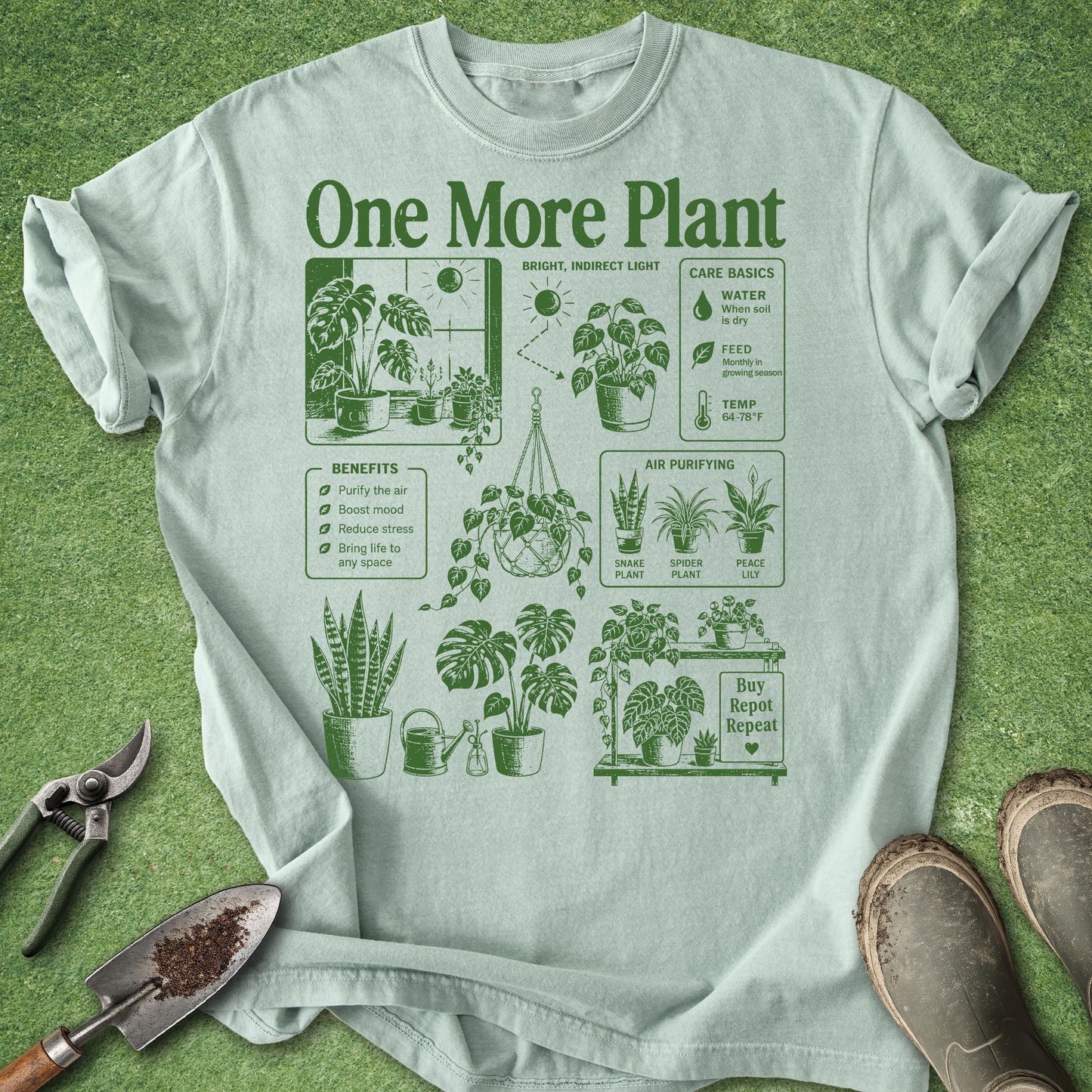 A light sage green graphic t-shirt laid flat on a green grass-like surface, featuring a dark green 'One More Plant' gardening infographic. Gardening props including pruning shears, a trowel with soil, and work boots are visible around the shirt.