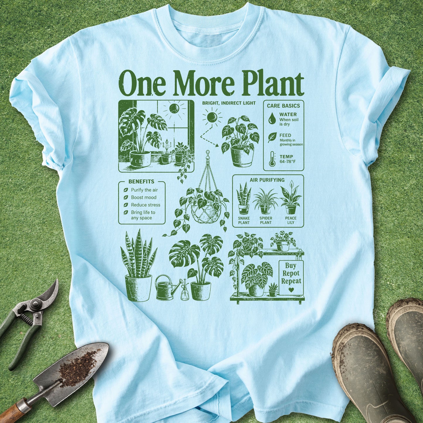 A light blue graphic t-shirt with a green 'One More Plant' design, laid flat on a green grass-like surface with gardening props including pruning shears, a trowel, and boots.