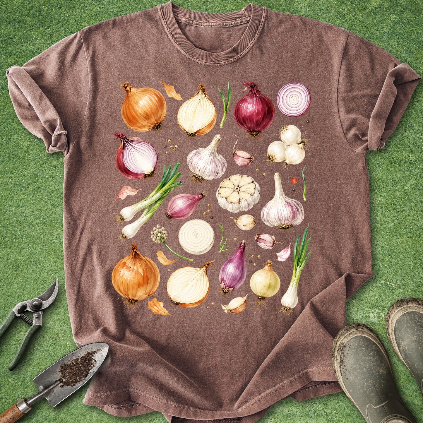 A brown short-sleeve crewneck t-shirt laid flat on green grass, featuring a colorful graphic print of various vegetables like onions, garlic, and scallions. Gardening tools and boots are styled around the shirt.