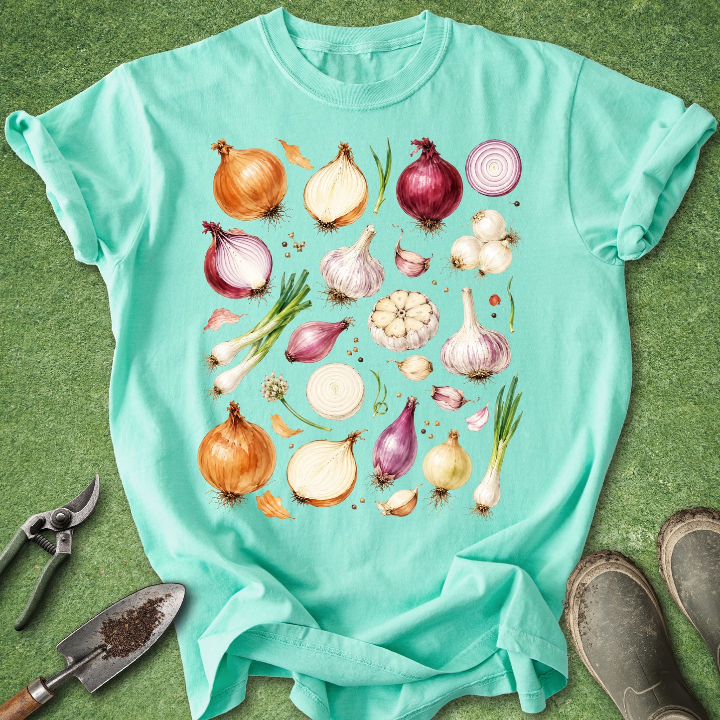 A mint green graphic t-shirt laid flat on a green grass-like surface, featuring a watercolor-style print of various onions, garlic, and herbs. A gardening trowel is visible on the bottom left, and a pair of muddy work boots is on the bottom right.