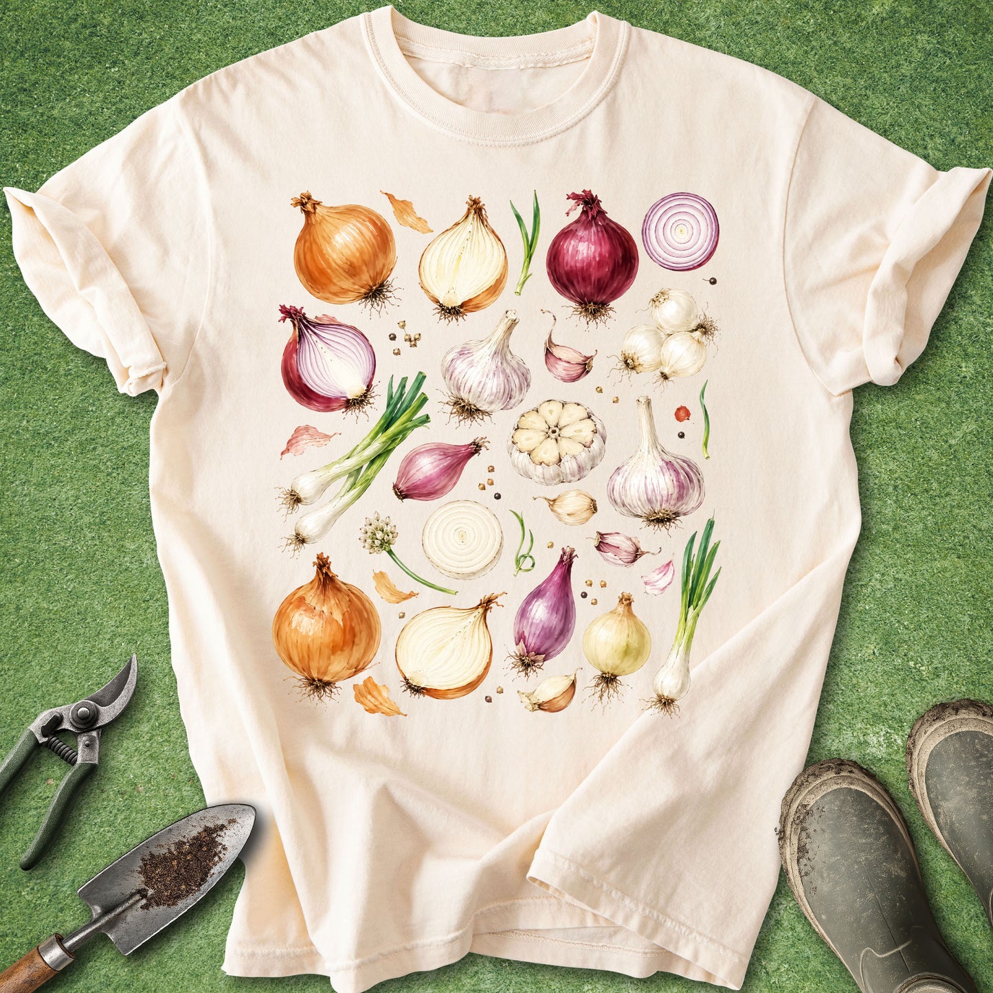 A cream-colored short-sleeve t-shirt with a watercolor-style print of various garden vegetables (onions, garlic, scallions) arranged on a green grass-like background. Gardening props, including a trowel, pruning shears, and boots, are visible around the shirt.