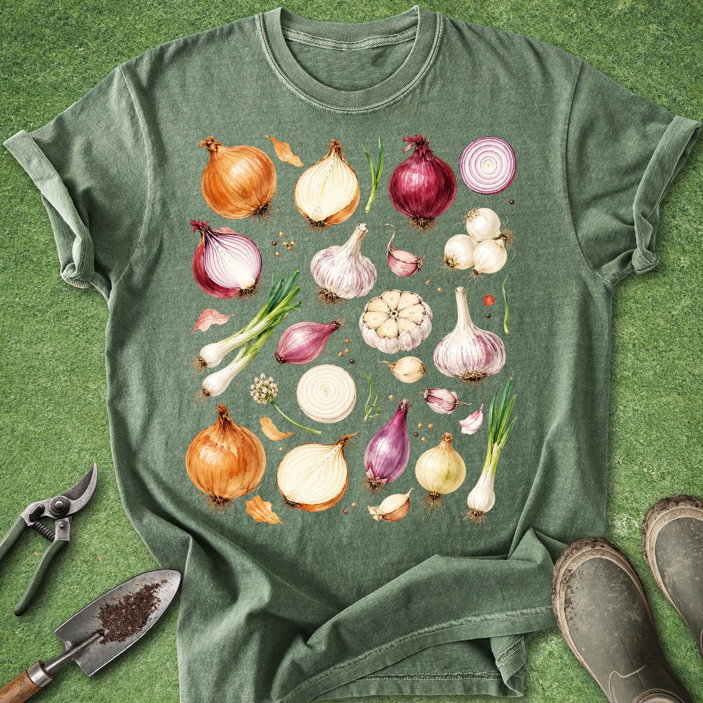 A green short-sleeve t-shirt laid flat on a grass-like surface, featuring a large printed graphic of various onions, garlic, and scallions in watercolor style. Gardening props, including pruning shears, a trowel, and boots, are visible around the shirt.