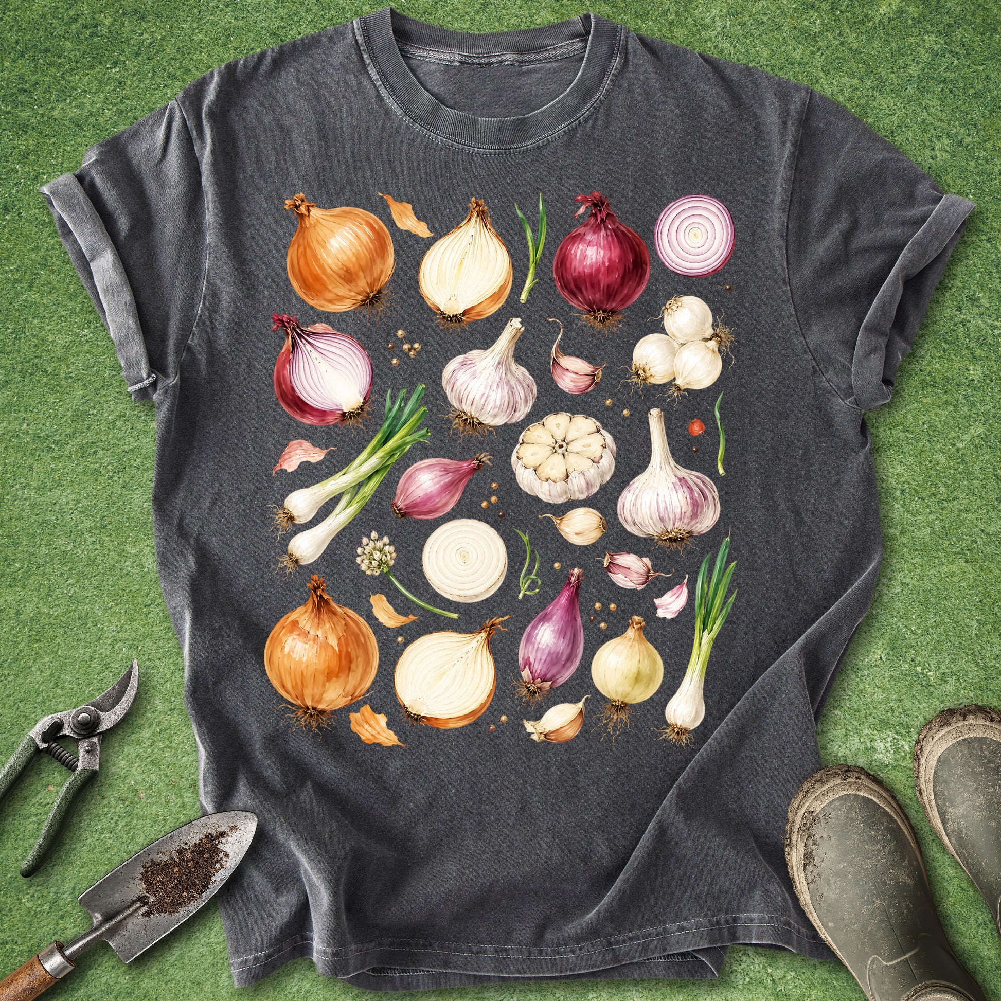 A dark charcoal gray t-shirt featuring a colorful watercolor-style graphic of various garden vegetables (onions, garlic, scallions, shallots) arranged in a grid. The t-shirt is styled in a flat-lay on green grass with gardening tools (trowel, pruning shears) and boots visible around the edges.