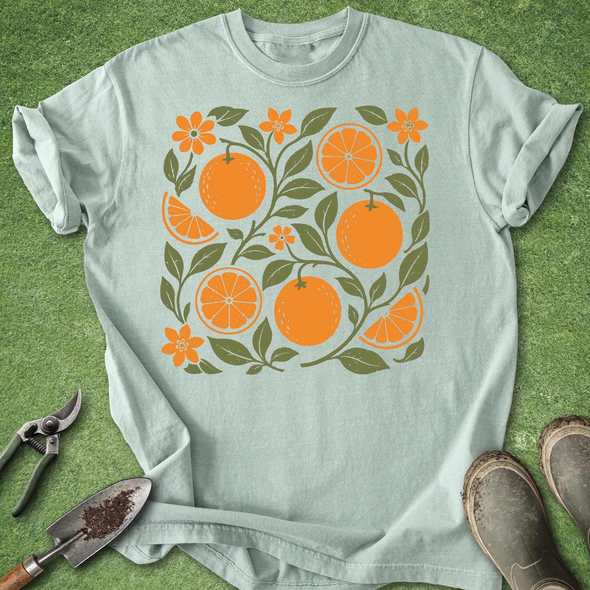 A light sage green t-shirt with a graphic print of oranges, orange slices, and green leaves, styled flat on a green grass background with gardening tools and boots.