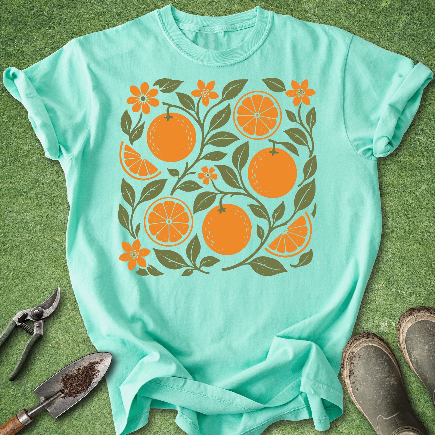A mint green short-sleeve t-shirt with a graphic print of oranges, orange slices, and leaves, laid flat on green grass with gardening tools and boots nearby.