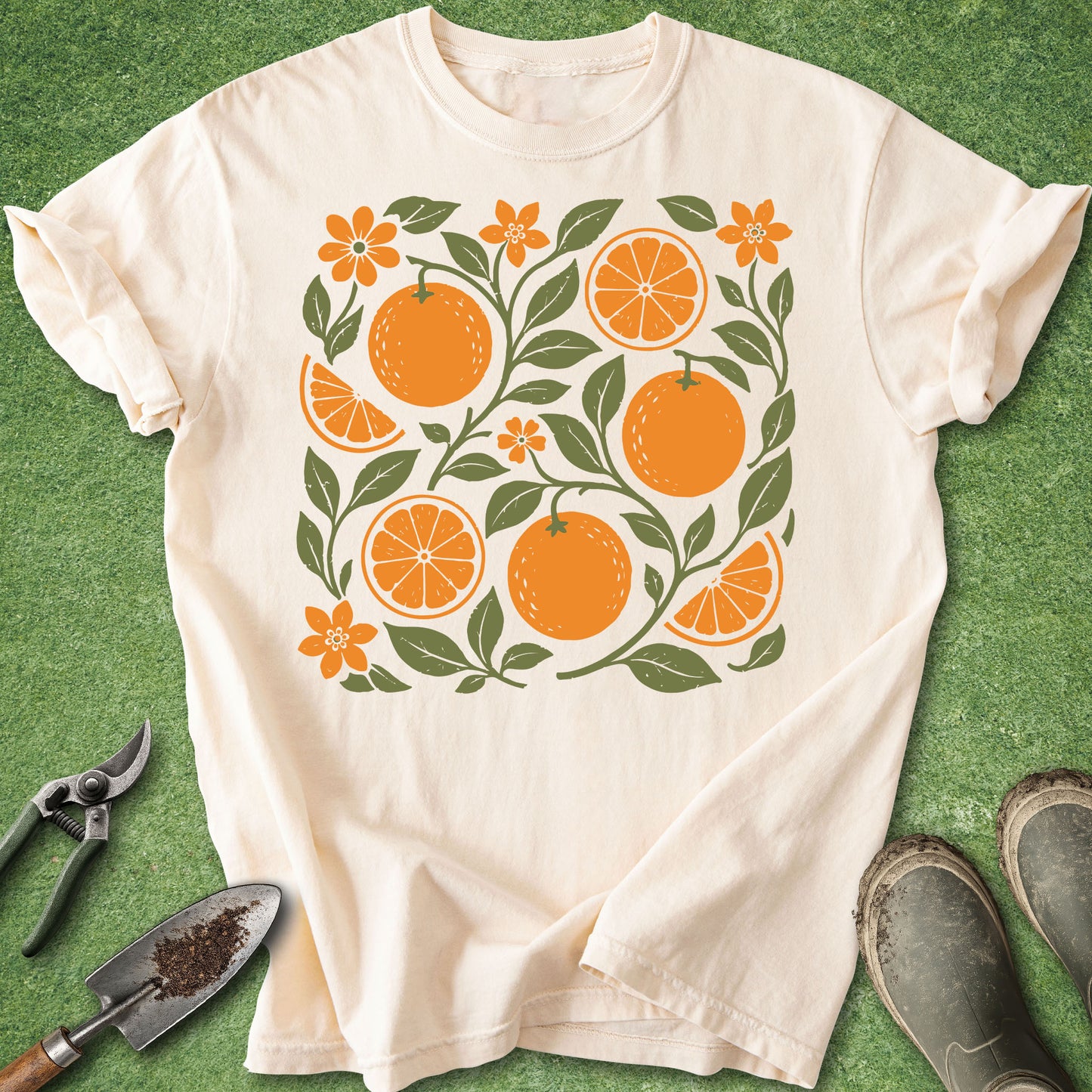 An off-white short-sleeve t-shirt with a retro-style orange and floral graphic print, laid flat on green grass with gardening tools and boots nearby.
