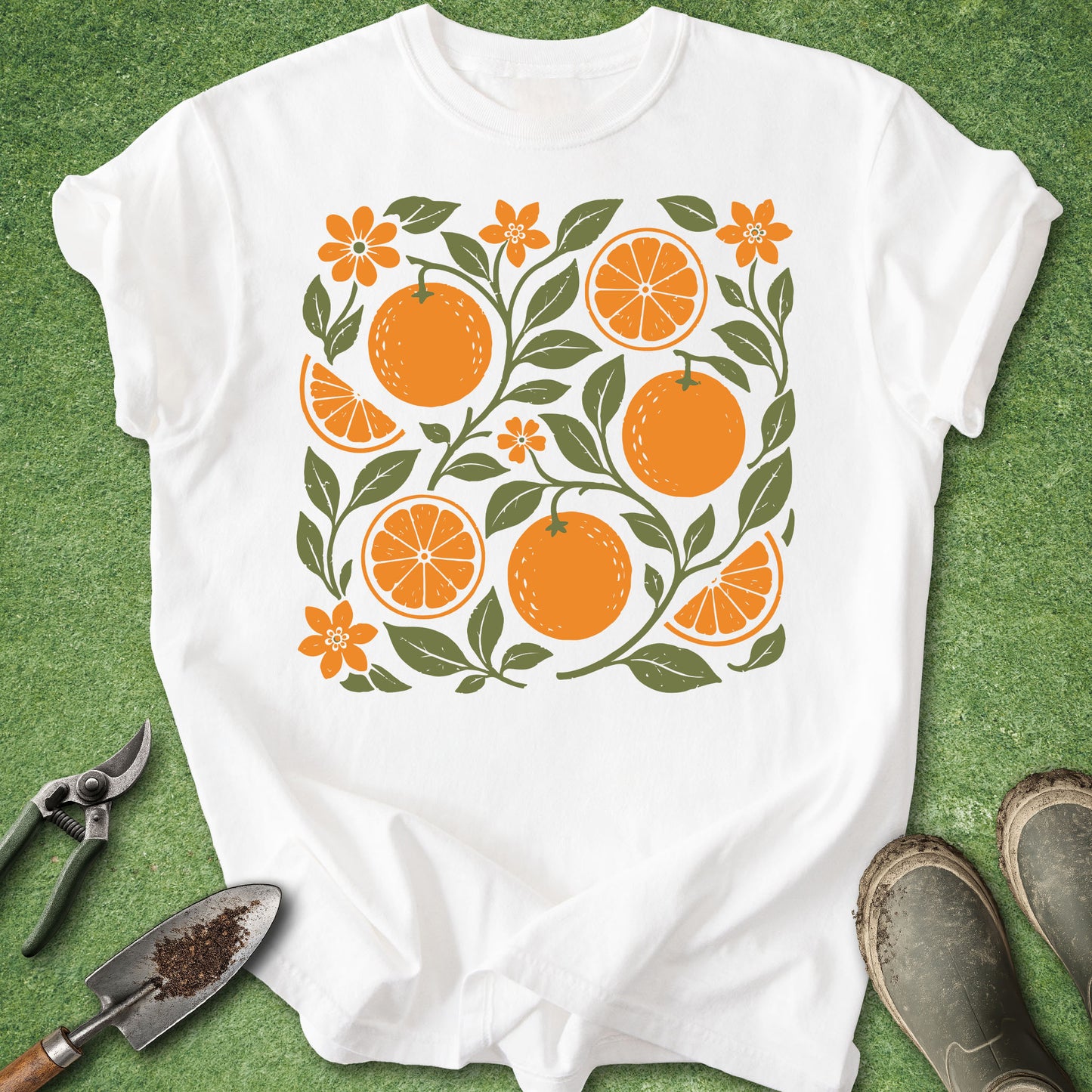 A white short-sleeve crewneck t-shirt laid flat on a green grass-like surface, featuring a central graphic of oranges, orange slices, and orange flowers with green leaves. Gardening tools and muddy boots are placed around the shirt as styling props.