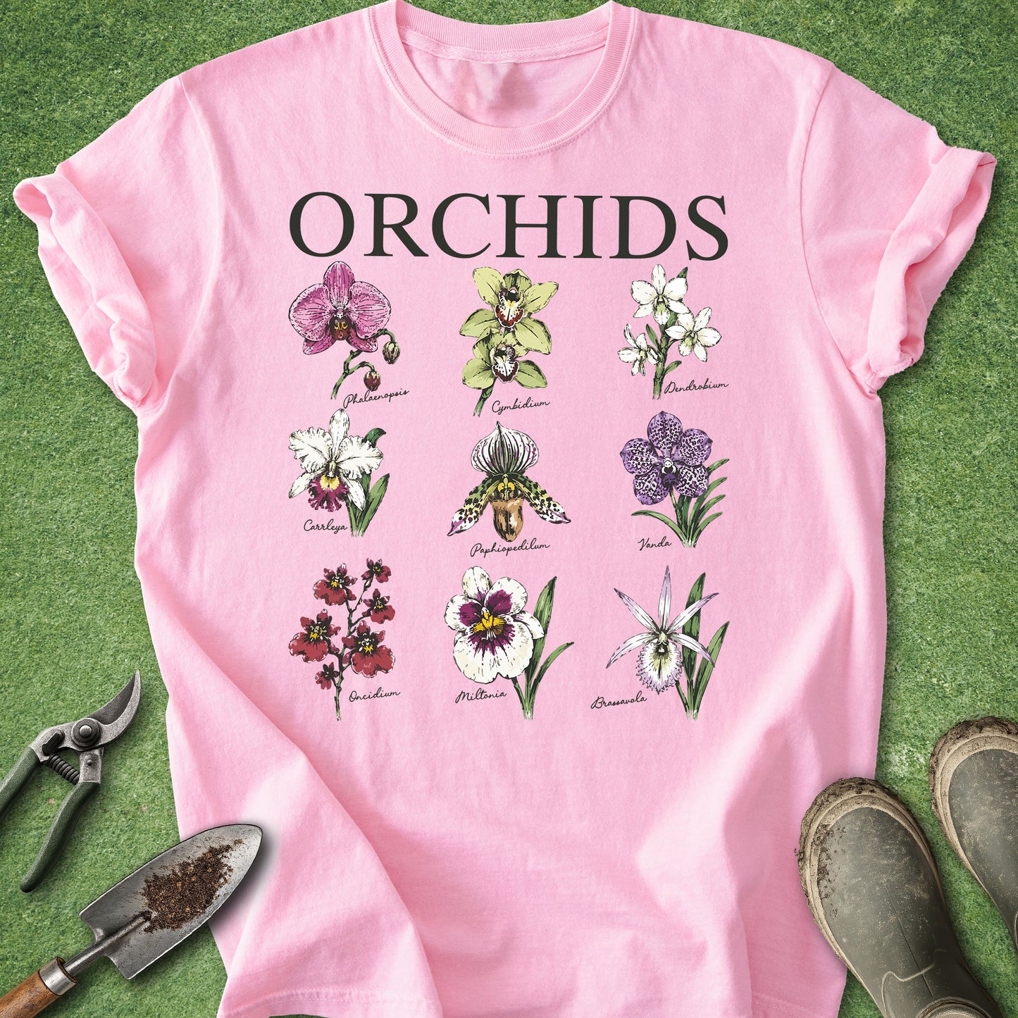 A pink short-sleeve T-shirt laid flat on green grass, featuring the word 'ORCHIDS' in large black letters above a grid of illustrated orchid flowers. Gardening tools and boots are partially visible around the shirt as styling props.