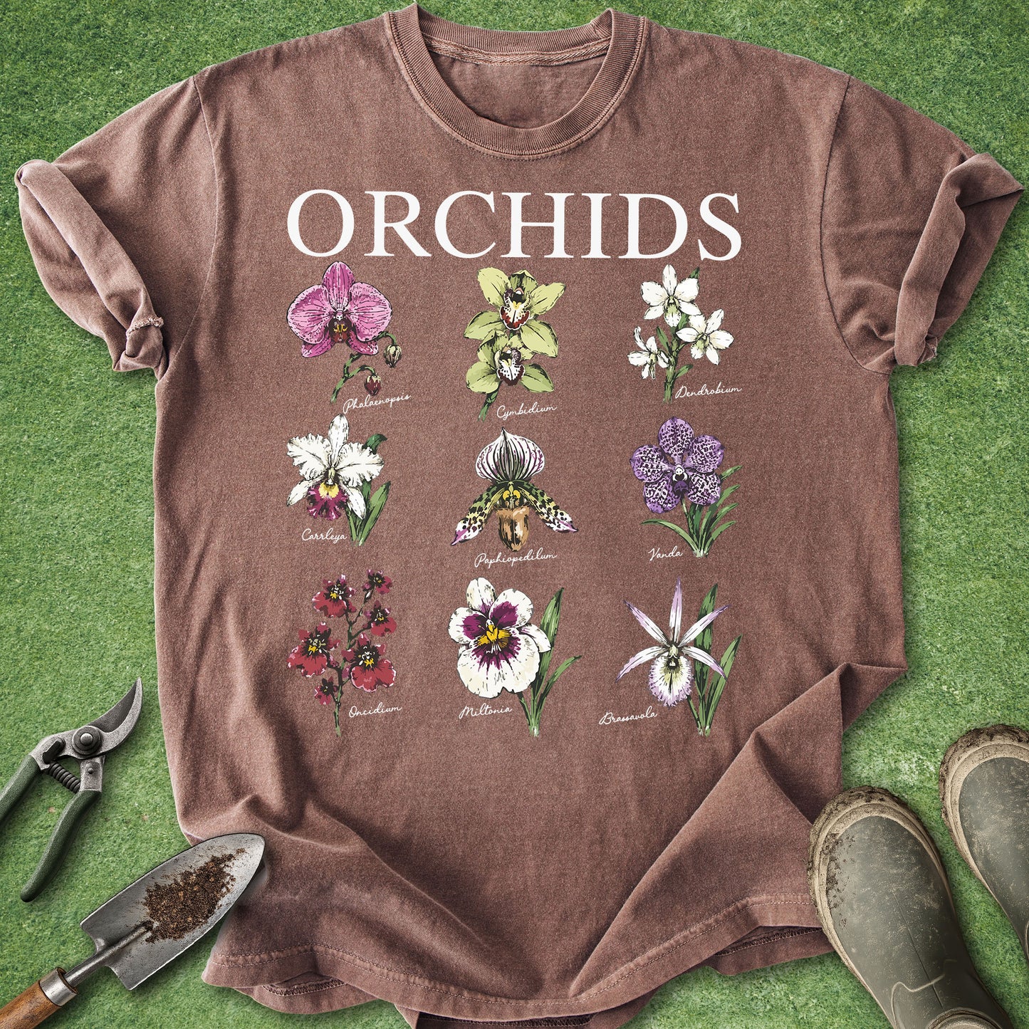 A brown short-sleeve t-shirt laid flat on green grass, featuring a white 'ORCHIDS' text print above a grid of ten different orchid and bulb illustrations. Gardening tools (trowel, pruning shears) and boots are placed around the shirt as props.