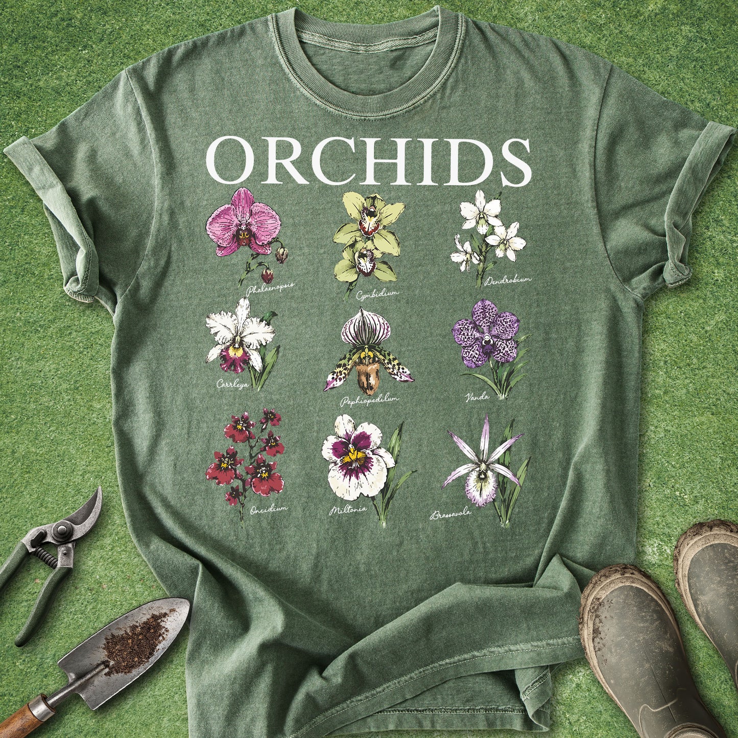 An olive green graphic t-shirt laid flat on a green grass-like surface, featuring the word 'ORCHIDS' in white above a grid of ten different orchid illustrations. Gardening tools (trowel, pruning shears) and boots are partially visible around the edges as styling props.