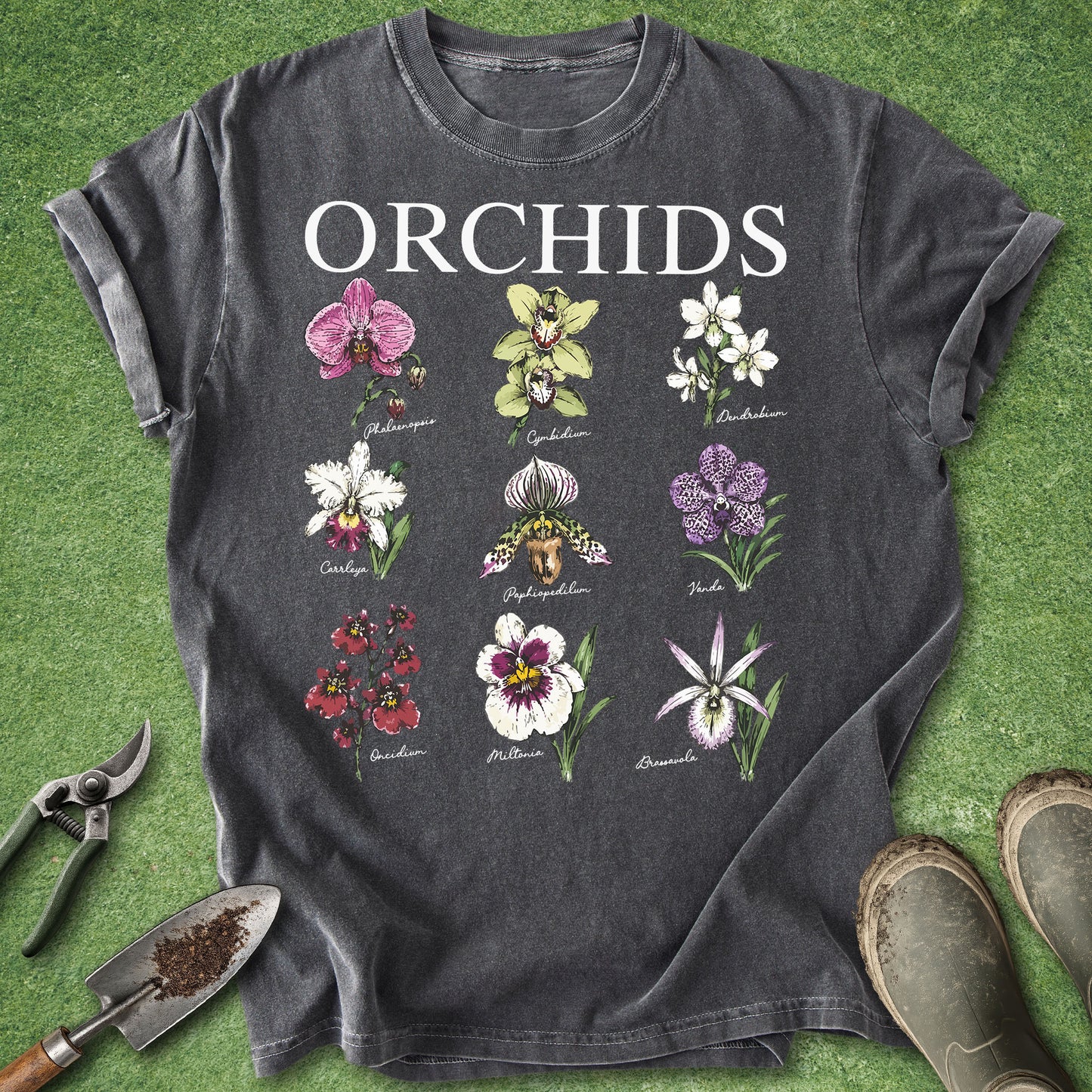 A dark charcoal gray t-shirt featuring a botanical graphic of various orchids with the word 'ORCHIDS' printed in white across the chest. The shirt is styled in a flat-lay arrangement on artificial green grass, surrounded by gardening tools (trowel, pruning shears) and gardening boots.