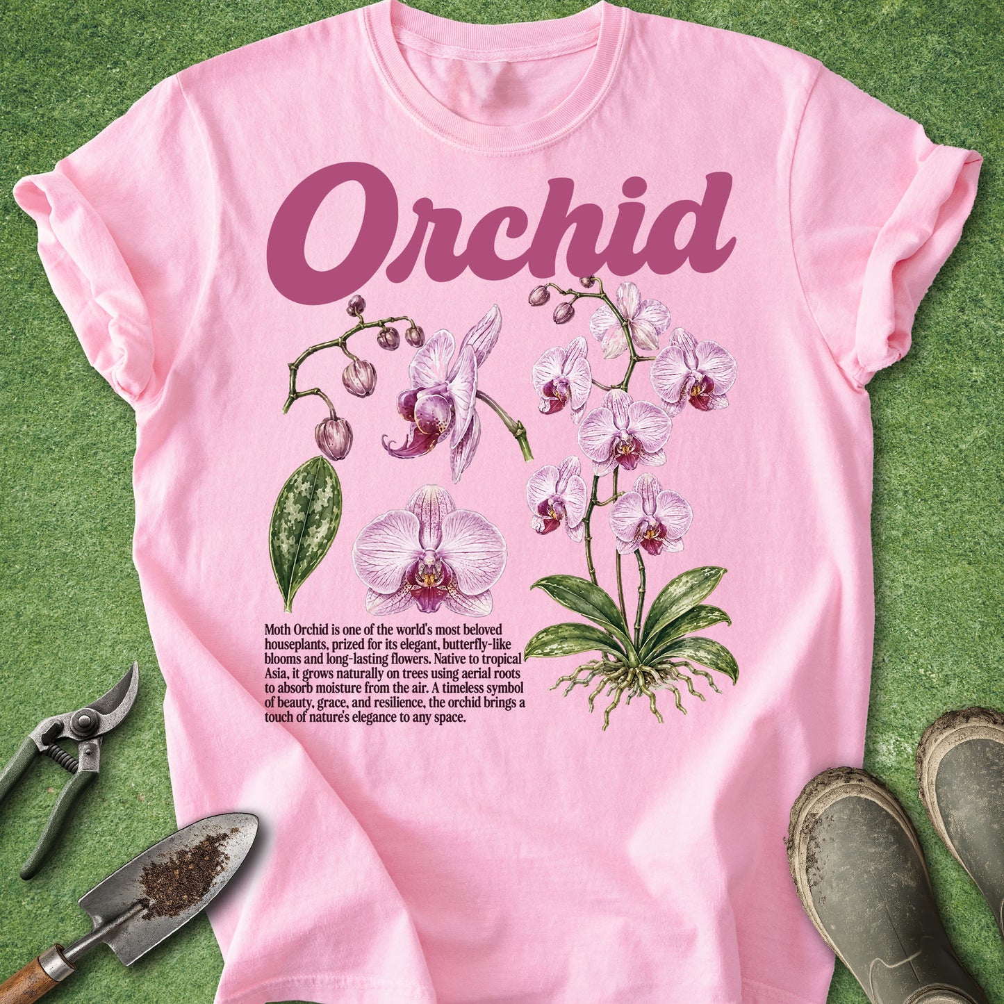A pink short-sleeve T-shirt laid flat on green grass-like background, featuring a large 'Orchid' wordmark above illustrated orchid flowers and a small text block. Gardening props including pruning shears, a trowel, and boots are placed around the shirt.