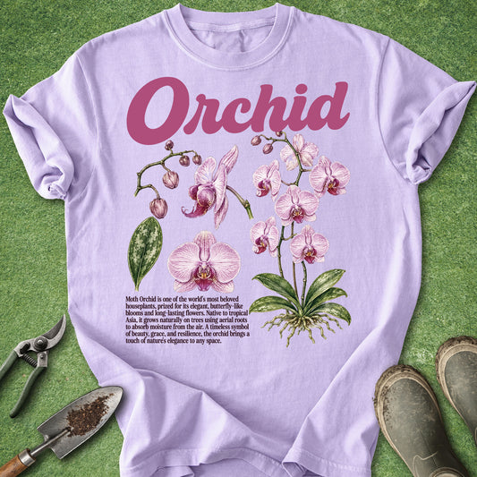 A light lavender graphic T-shirt laid flat on a green textured surface, featuring the word 'Orchid' in pink script above an illustrated pink orchid with green leaves and a small text block. Gardening props including pliers, a trowel, and boots are placed around the shirt.