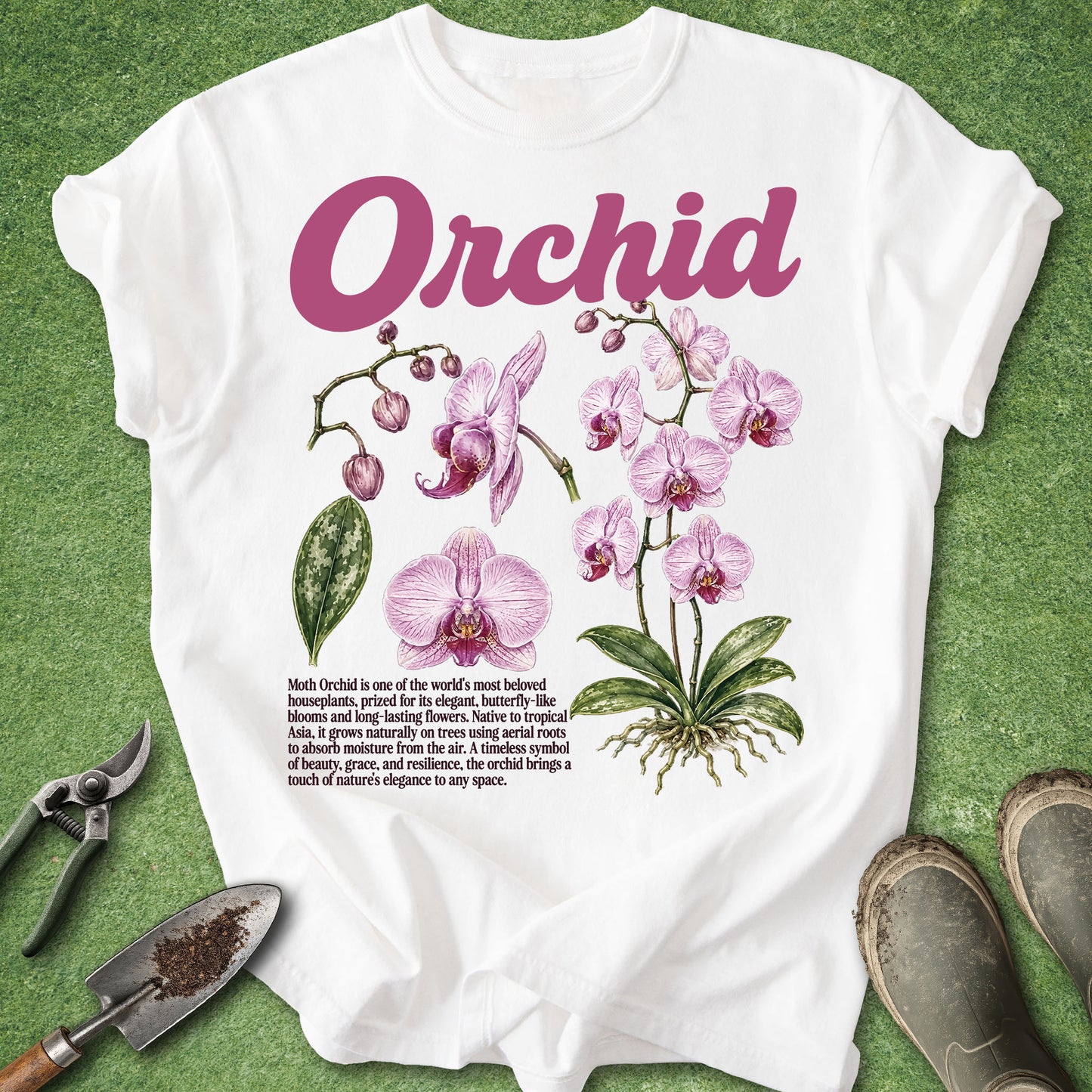 A white short-sleeve T-shirt laid flat on a green textured surface, featuring a large pink 'Orchid' title, pink orchid flowers, green leaves, and a small text block on the front. Gardening tools and boots are placed around the shirt as styling props.