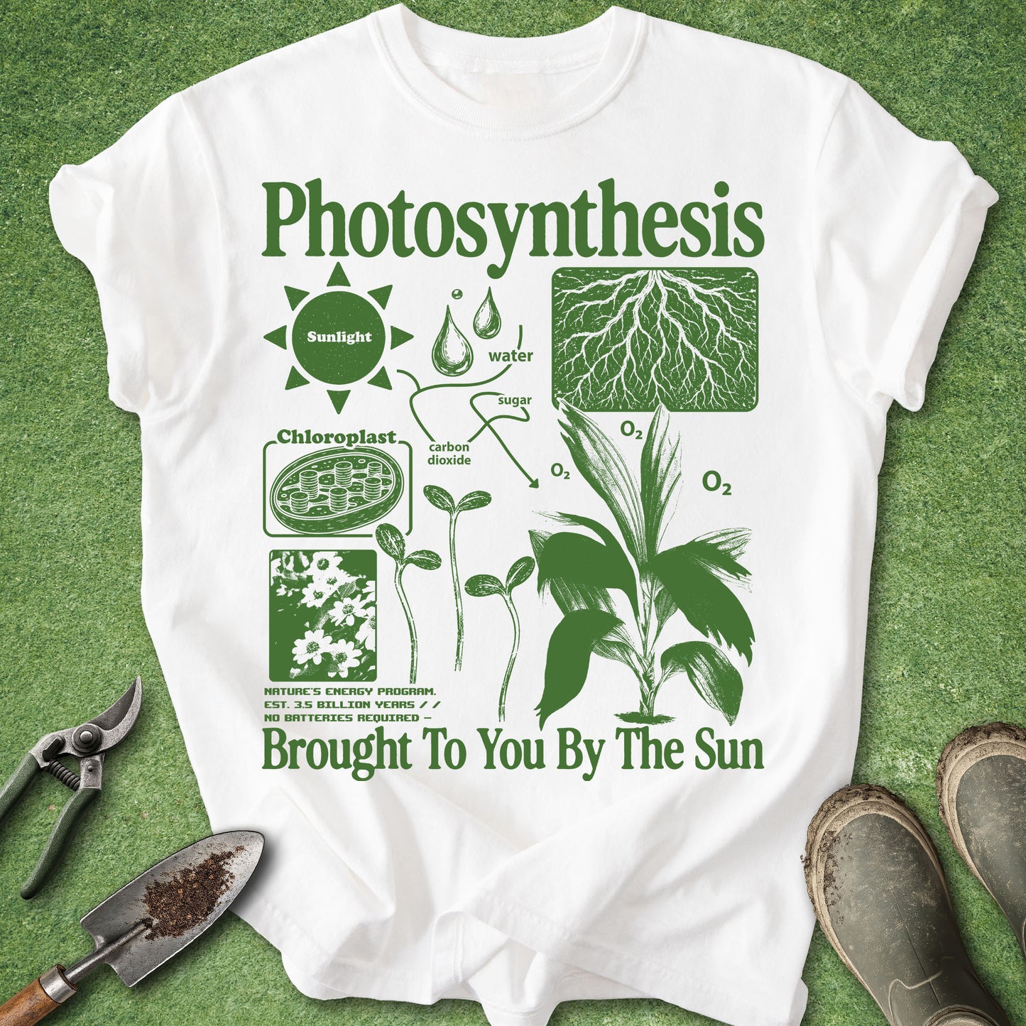 A white short-sleeve T-shirt laid flat on green grass, featuring a green printed diagram about photosynthesis with labels, leaves, roots, and the text 'Photosynthesis' and 'Brought To You By The Sun.' Gardening tools and boots are partially visible around the shirt as styled props.