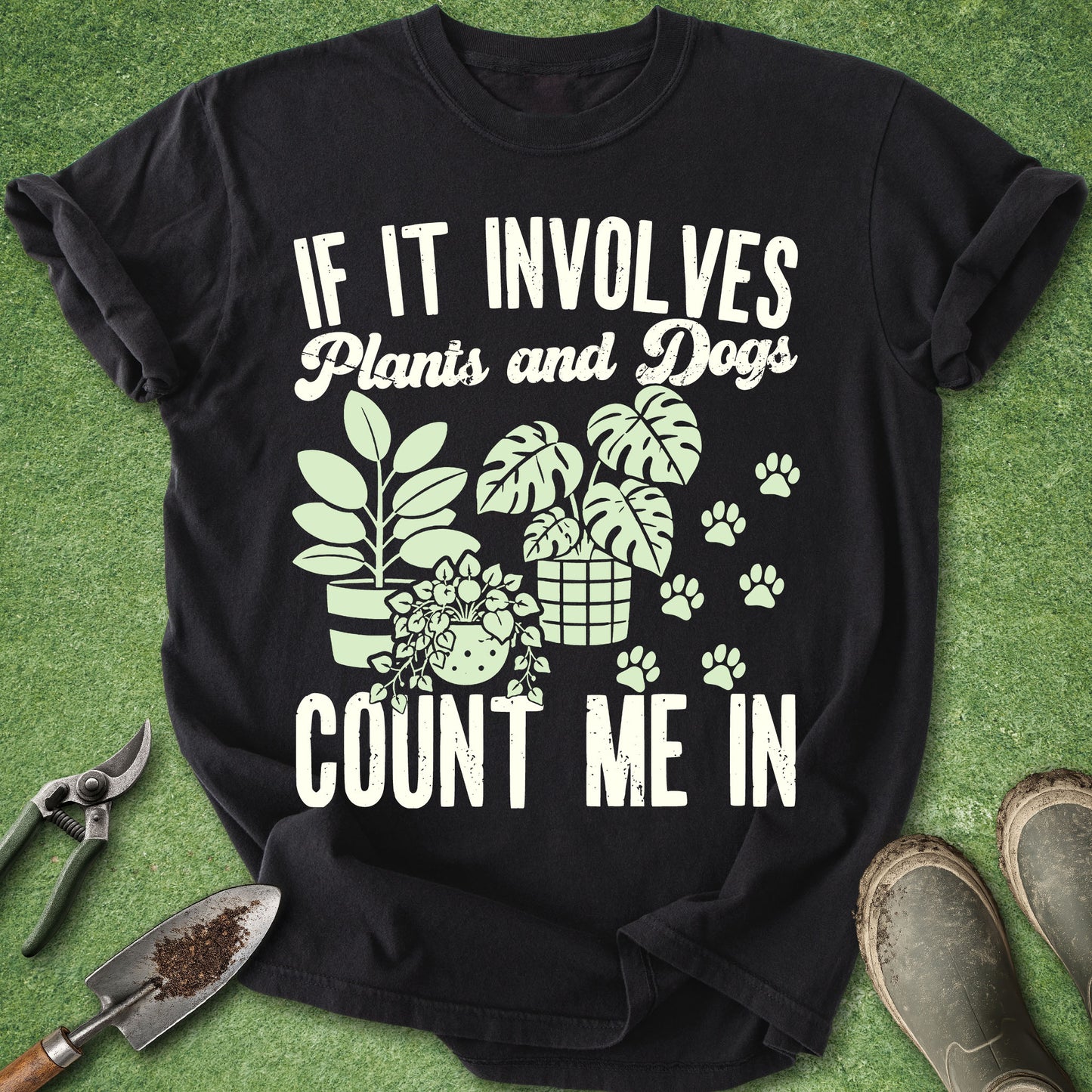 A black short-sleeve graphic T-shirt laid flat on green grass-like ground, with gardening tools and boots around it. The shirt has large white text reading 'IF IT INVOLVES Plants and Dogs COUNT ME IN' and a light green illustration of potted plants, leaves, and paw prints.